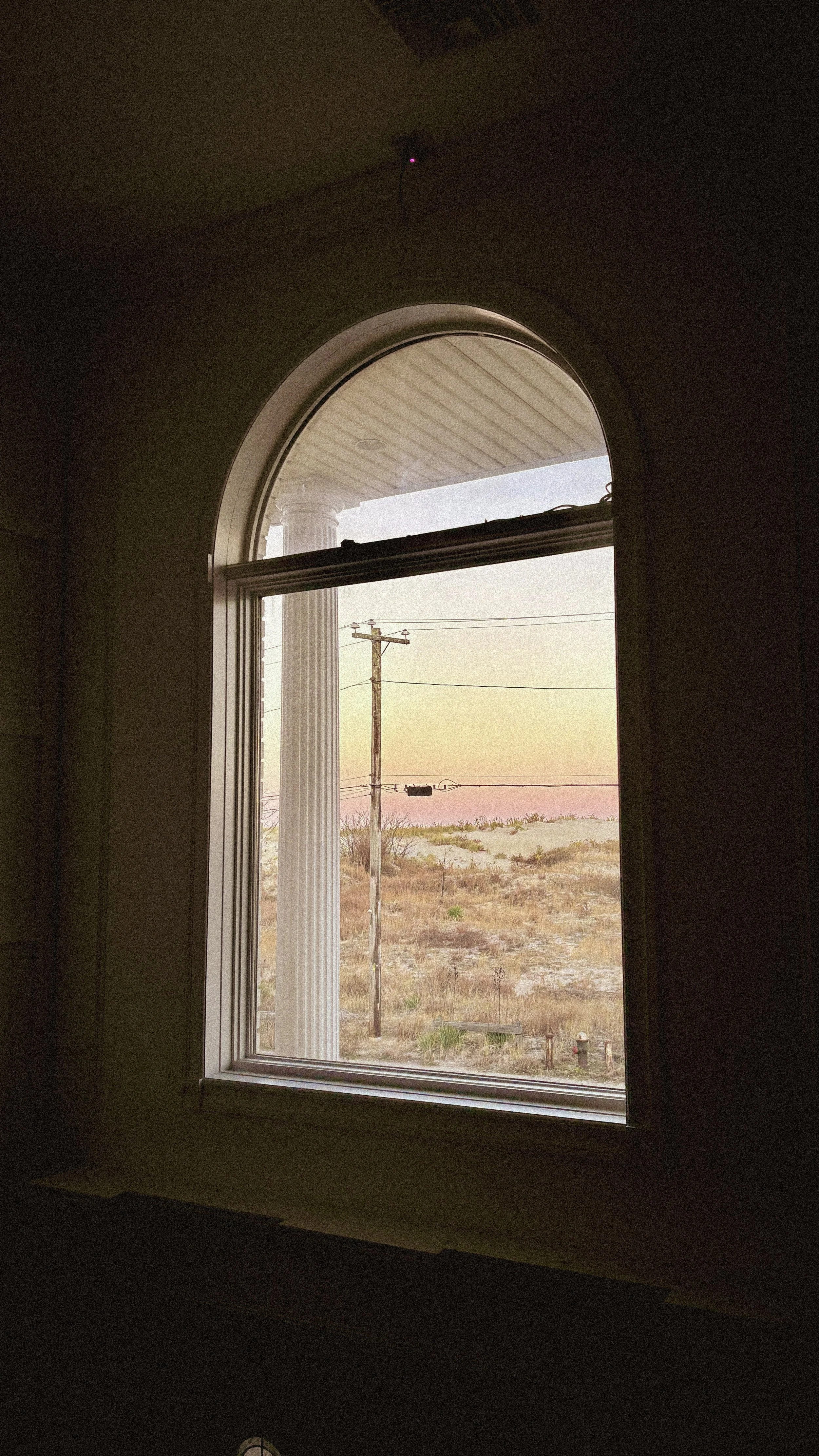 View from inside a building through a window showing a utility pole, a wheat field, and the sky at sunset or sunrise.