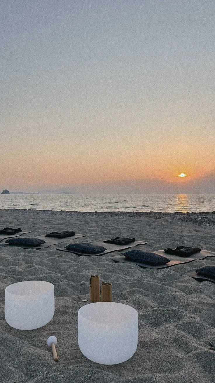 A beach scene at sunset with folded mats, ceramic singing bowls, and two wooden mallets on the sand.