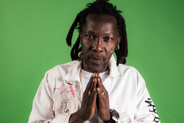 A man with dreadlocks and a beard, wearing a white jacket with pink and black writing, stands against a green background, holding his hands together in a prayer gesture.