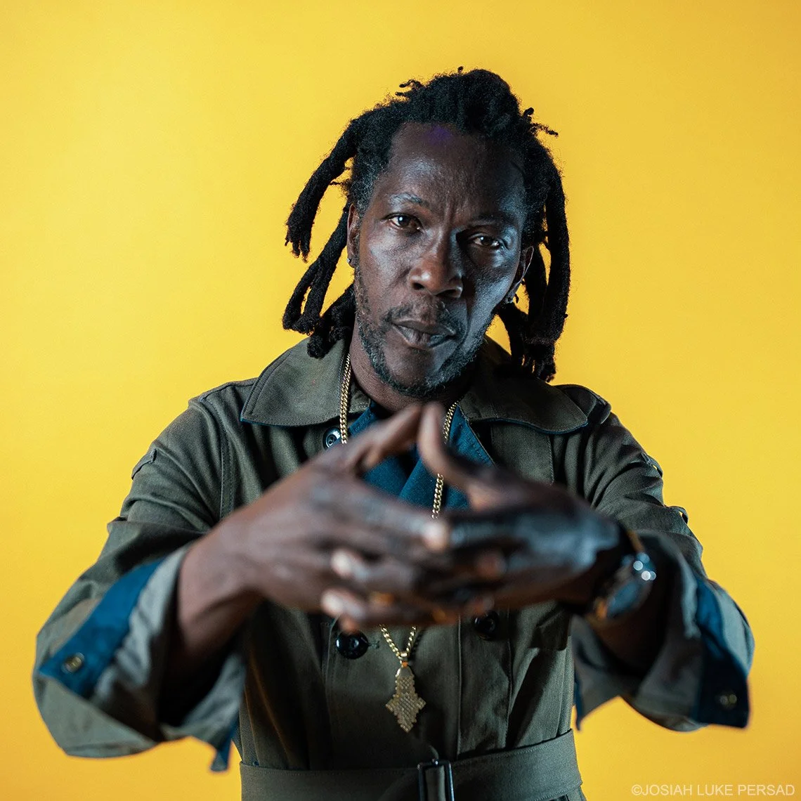 A man with dark skin and dreadlocks wearing a green jacket, a gold chain, and a crucifix necklace, poses against a solid yellow background. He is looking directly at the camera with a serious expression, with his hands clasped in front of him.