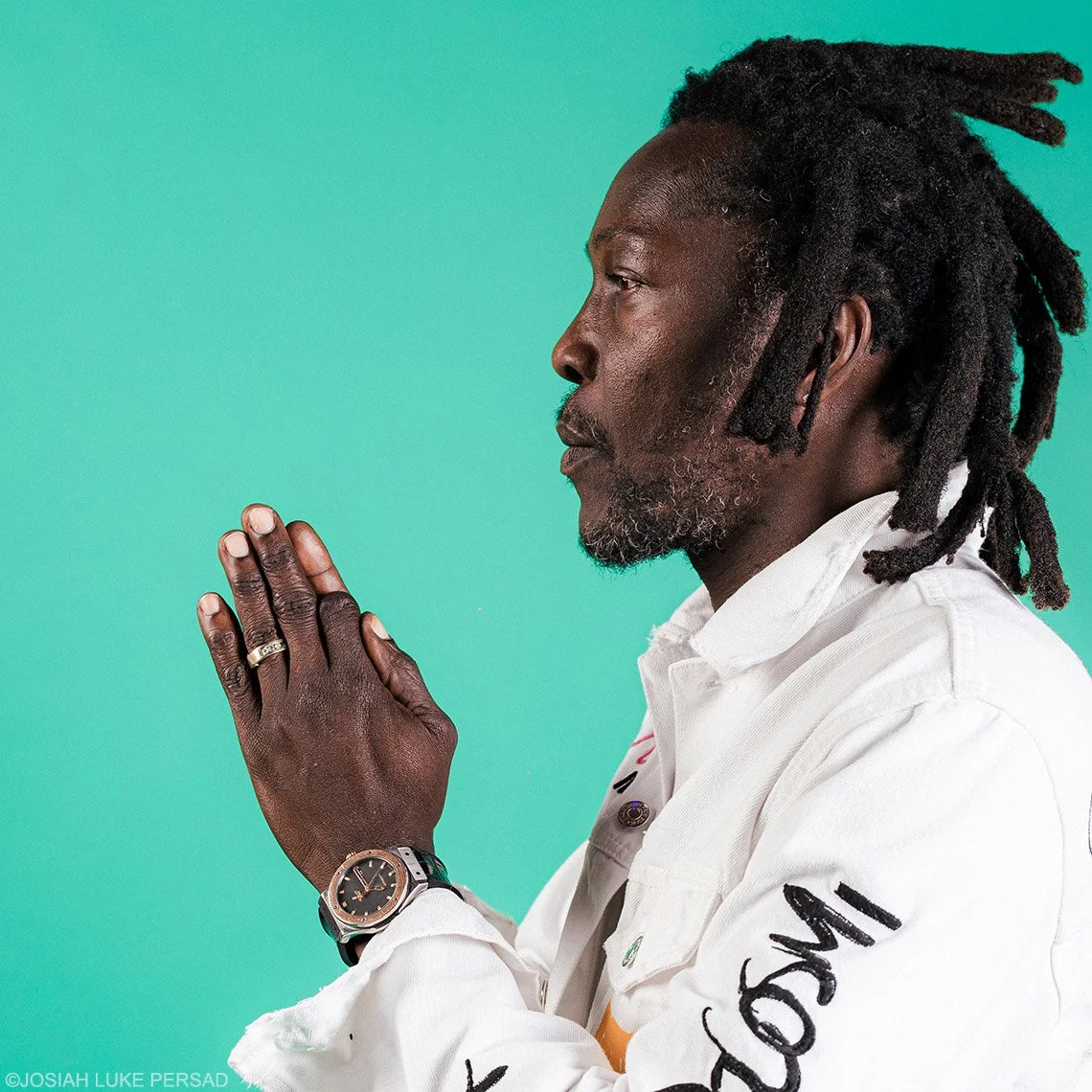 A man with dreadlocks, wearing a white jacket, holds his hands together in a prayer position against a teal background.