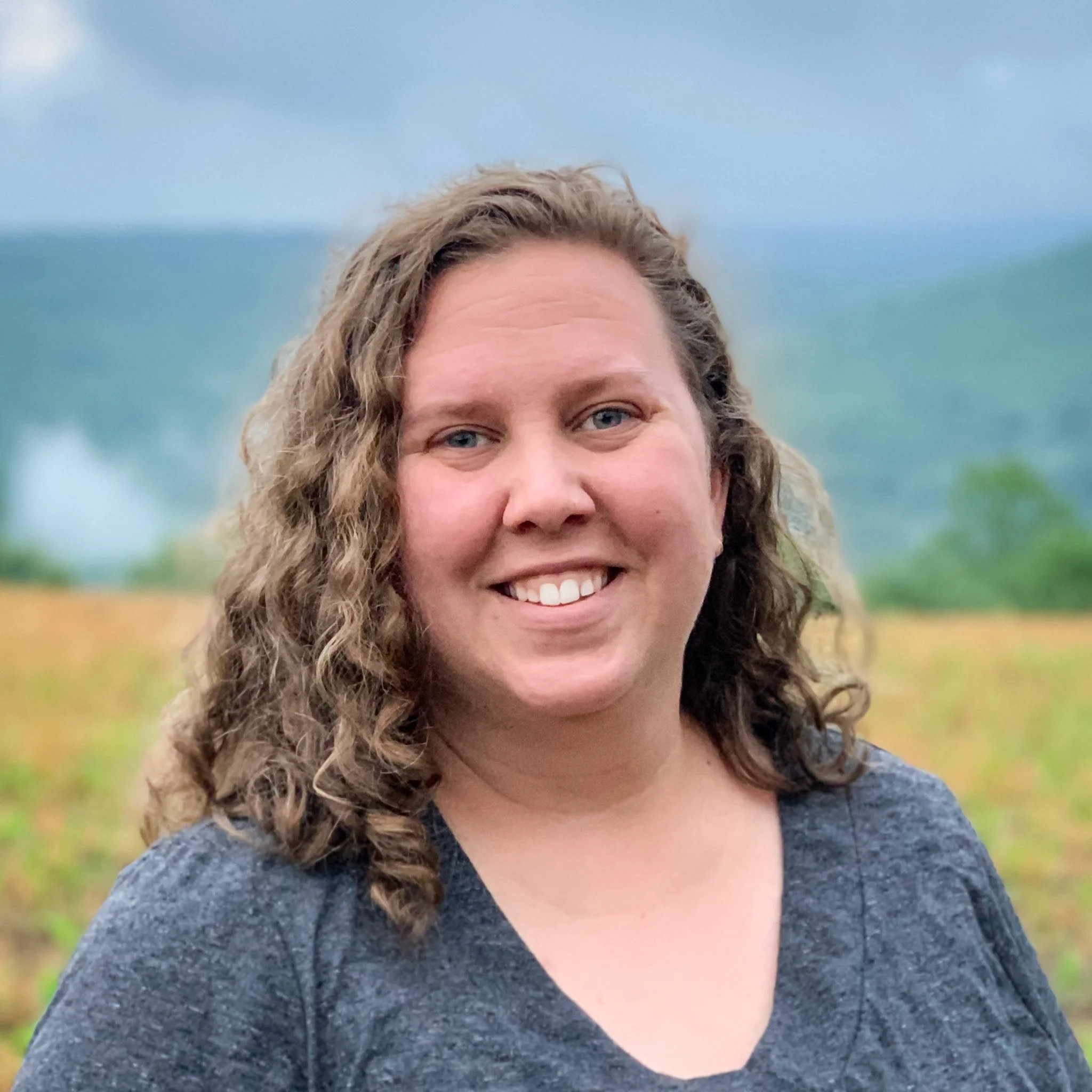 Close-up of a woman, Emily Hostetter, with curly brown hair, blue eyes, and smiling face, wearing a dark gray top, outdoors with a blurred background of blue sky and green grass.