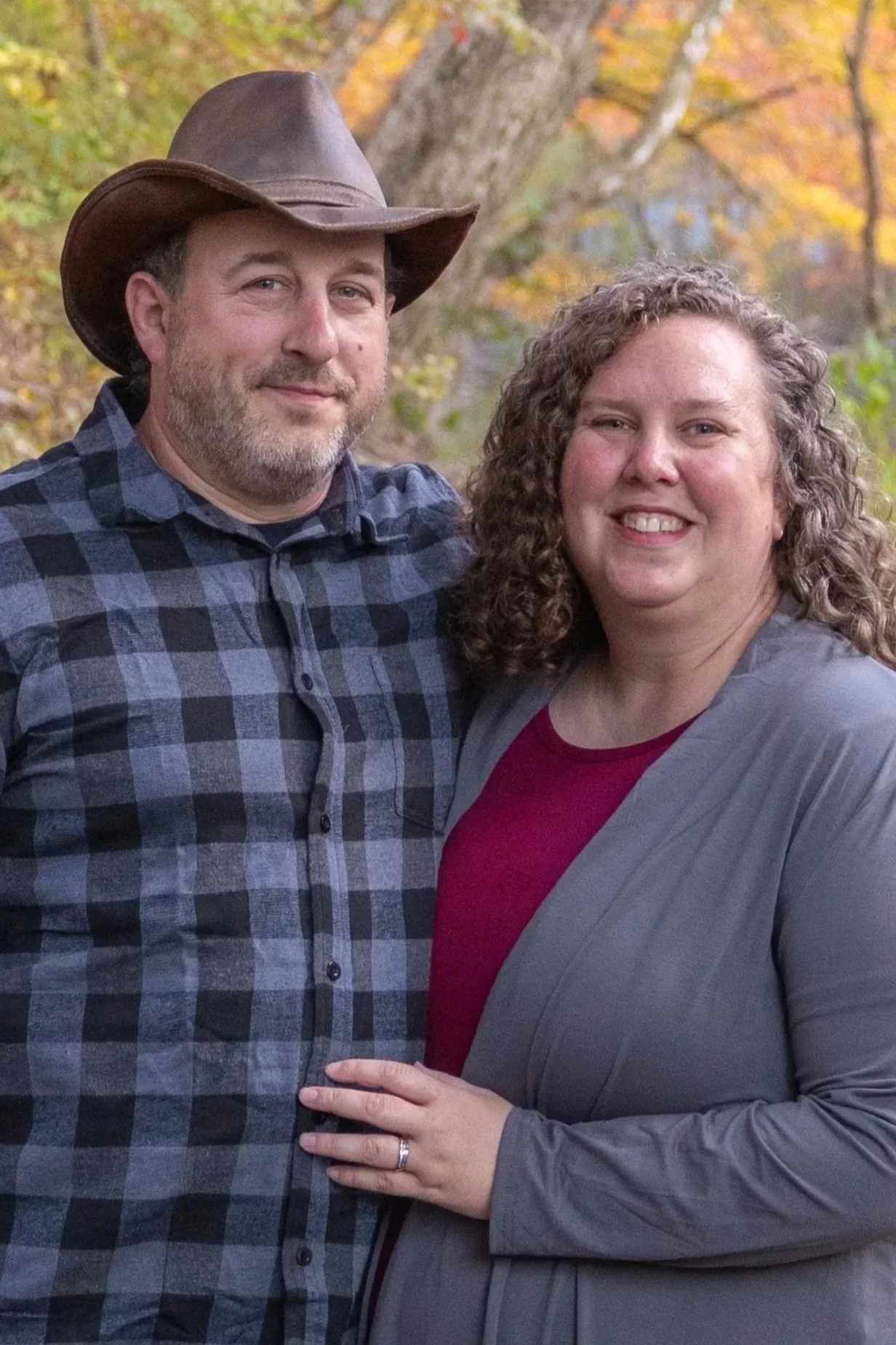 A man and woman standing outdoors during fall, smiling at the camera. The man is wearing a plaid shirt and a cowboy hat, while the woman has curly hair and is dressed in a gray blazer over a maroon top. There are trees with colorful autumn leaves in the background.
