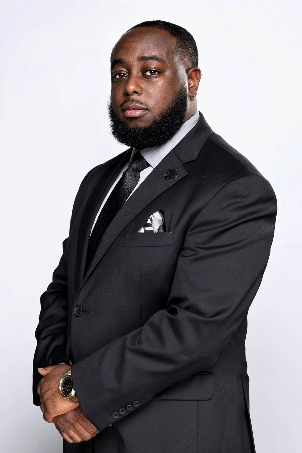 A man in a black suit, gray shirt, and black tie standing with arms crossed against a plain white background.