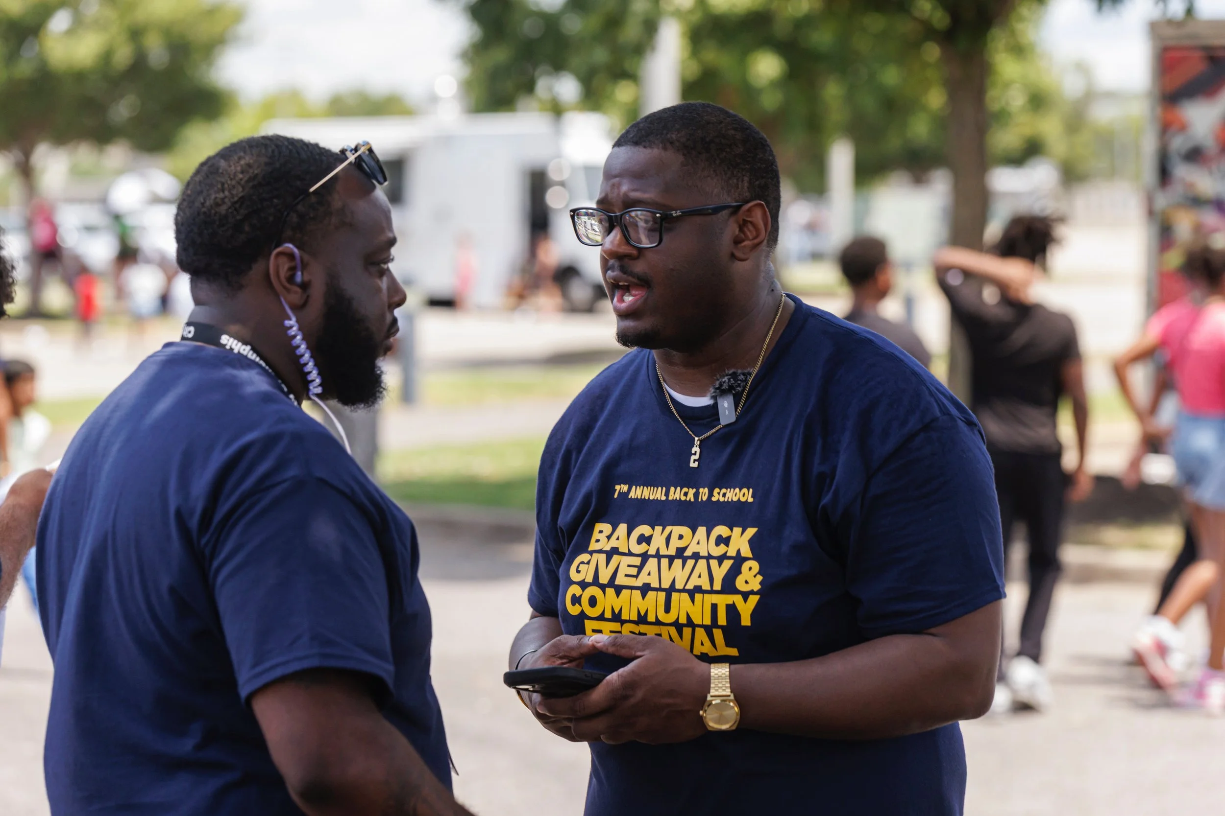 Two men talking at a community event, one wearing a dark blue T-shirt with yellow text about a backpack giveaway and school festival, outdoors with other people and trees in the background.