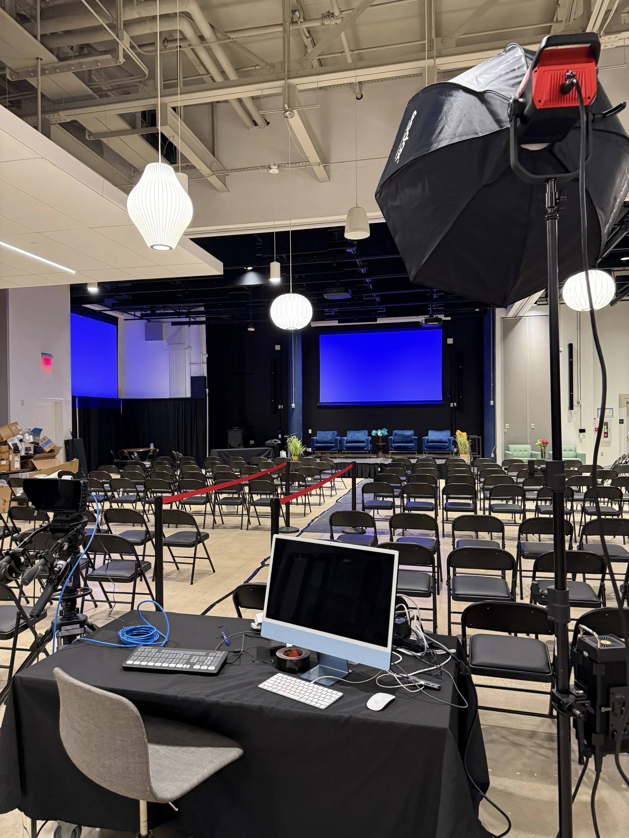 Empty conference room setup for a panel or presentation with a stage, large screens, rows of chairs, and professional lighting and camera equipment