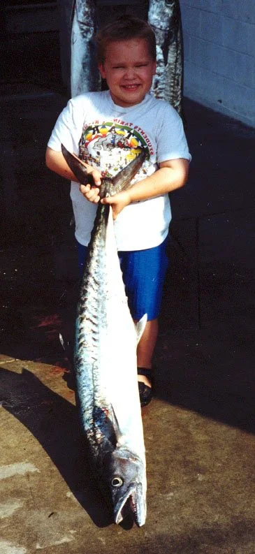A young boy with a big smile holding a large fish on the ground in front of him.