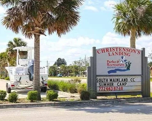 Sign for Christenson's Landing at South Mainland Community Center with a palm tree and a boat on a trailer in the background.