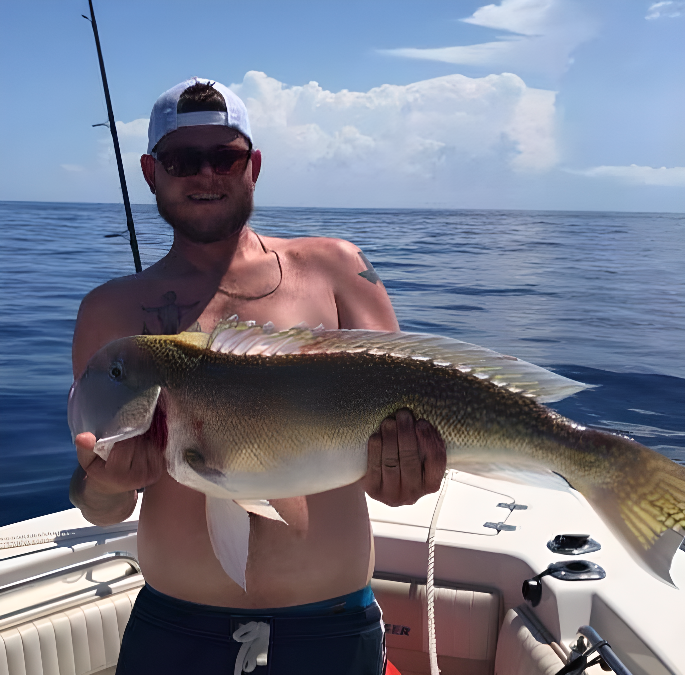 A man with sunglasses, a white cap backwards, and tattoos on his arms, shirtless, holding a large fish on a boat in open water with a cloudy sky in the background.