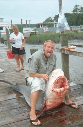 Man sitting on a dock holding a large fish with its mouth open, while a woman stands nearby with a red container, in a marina.