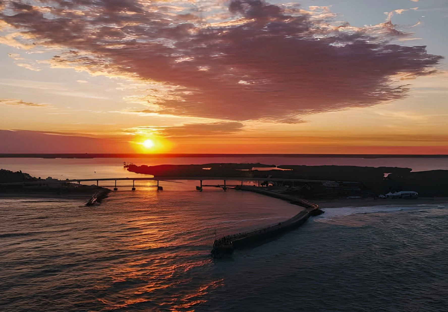 A sunset over a body of water with a bridge and land in the background, partially cloudy sky with pink and purple hues.