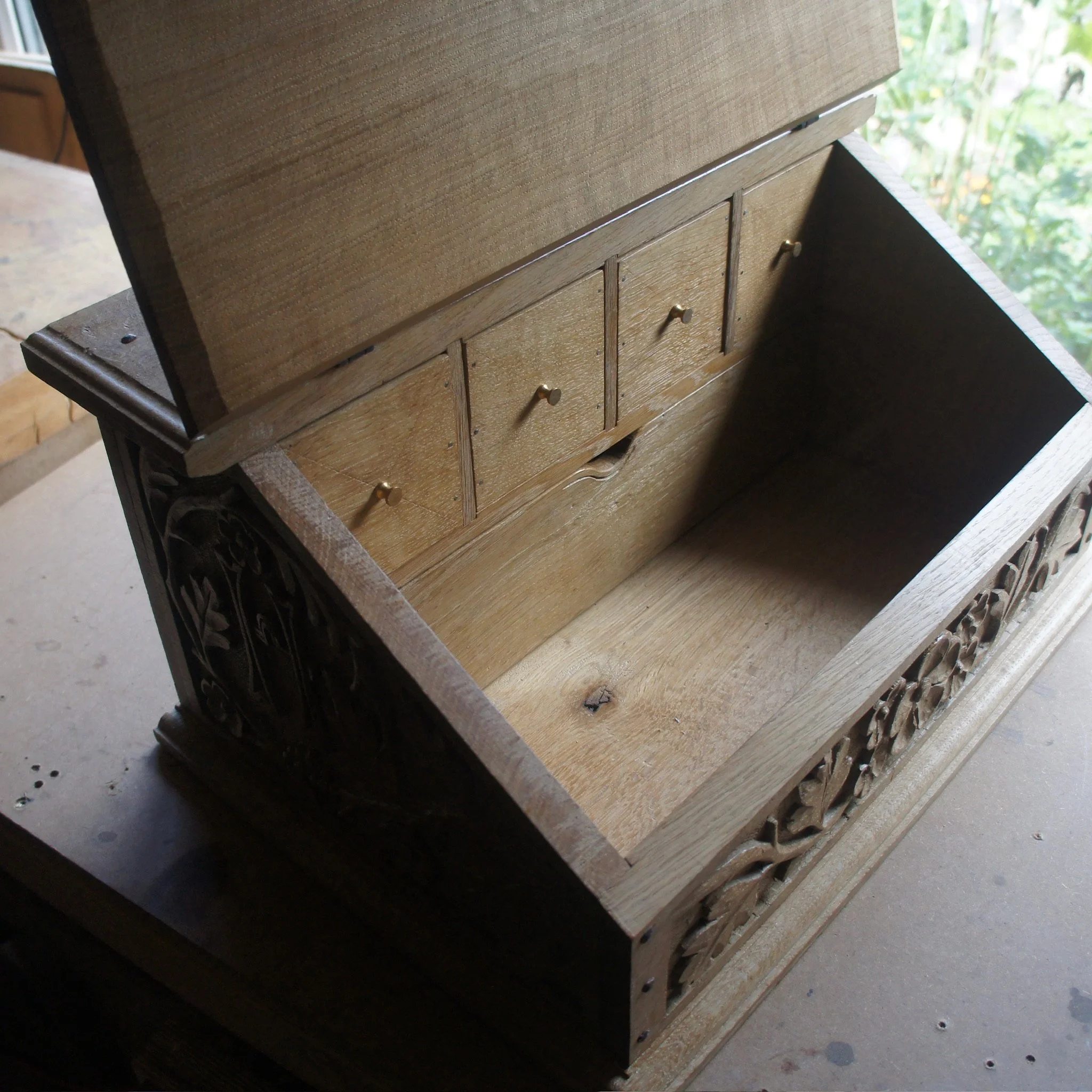 A wooden piece of furniture with drawers, placed on a surface with visible cracks and holes, with a background of green foliage outside a window.