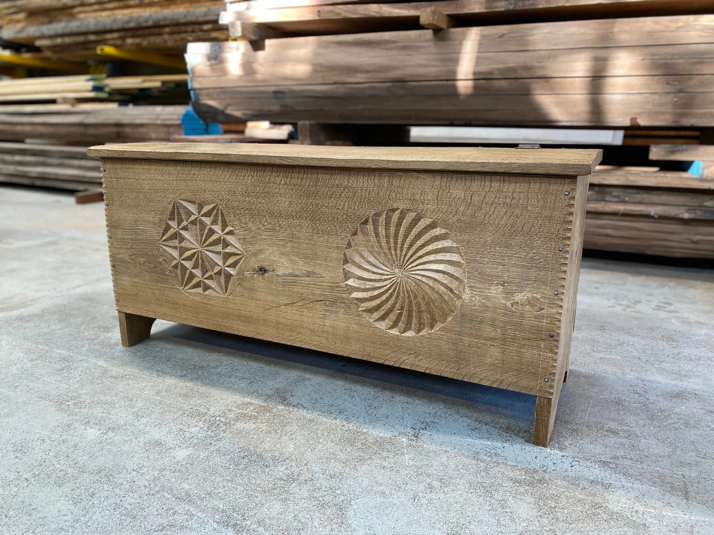 Wooden furniture piece, possibly a bench or chest, with carved geometric and spiral patterns on the side, placed outdoors with stacks of wood in the background.