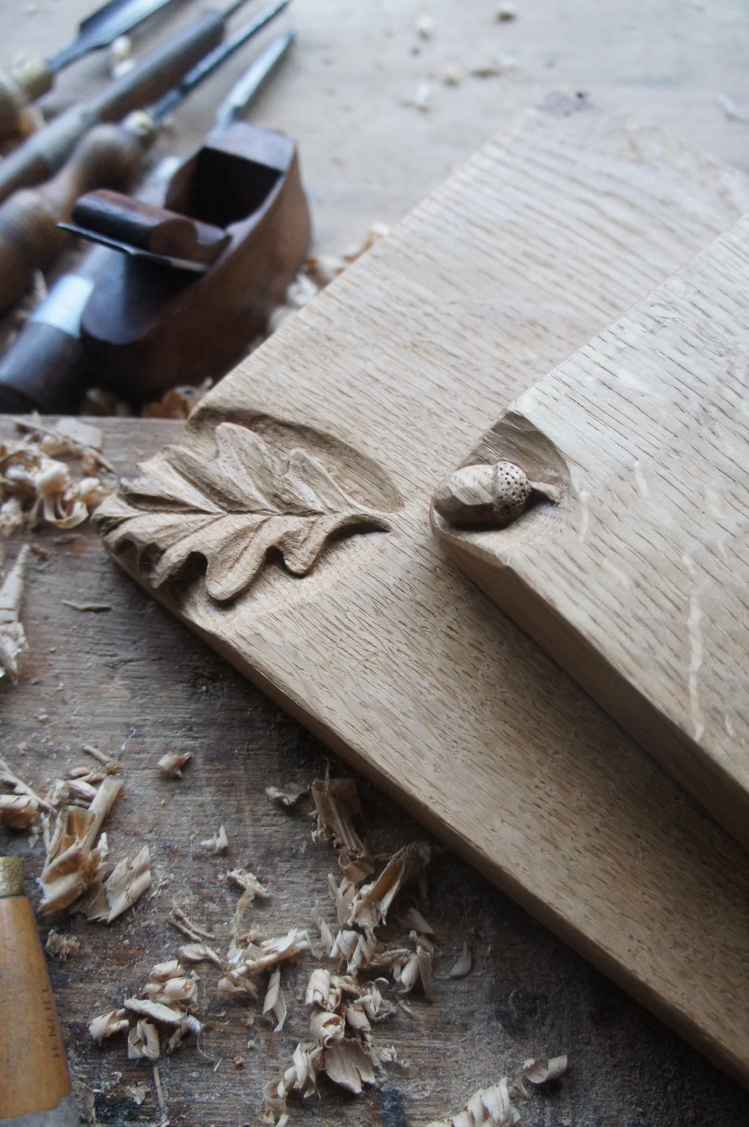 Close-up of wood shaving scraps and a carved wood piece on a workbench, with woodworking tools in the background.