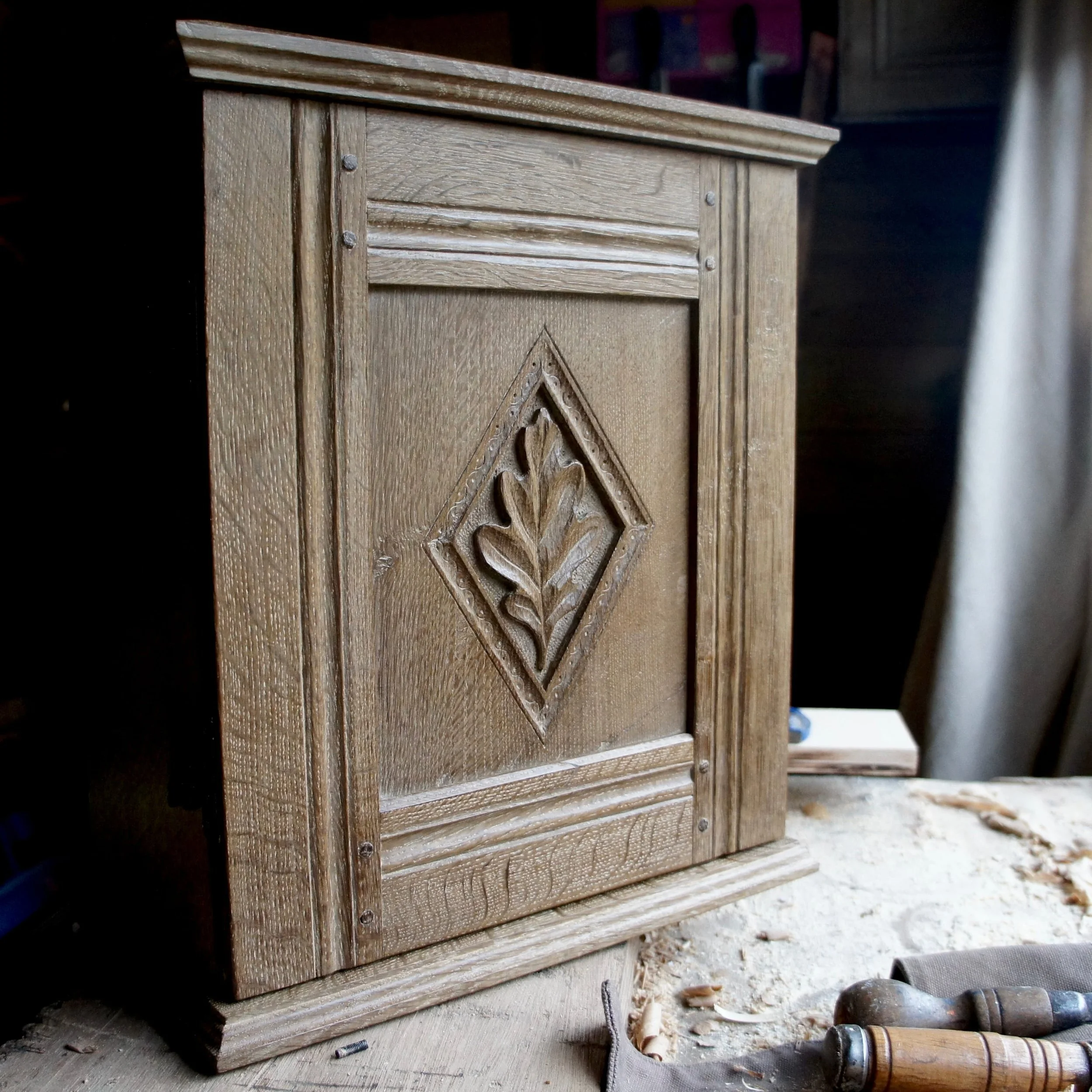 Close-up of a wooden furniture piece with decorative carvings and visible wood grain, placed on a dusty surface with tools nearby.