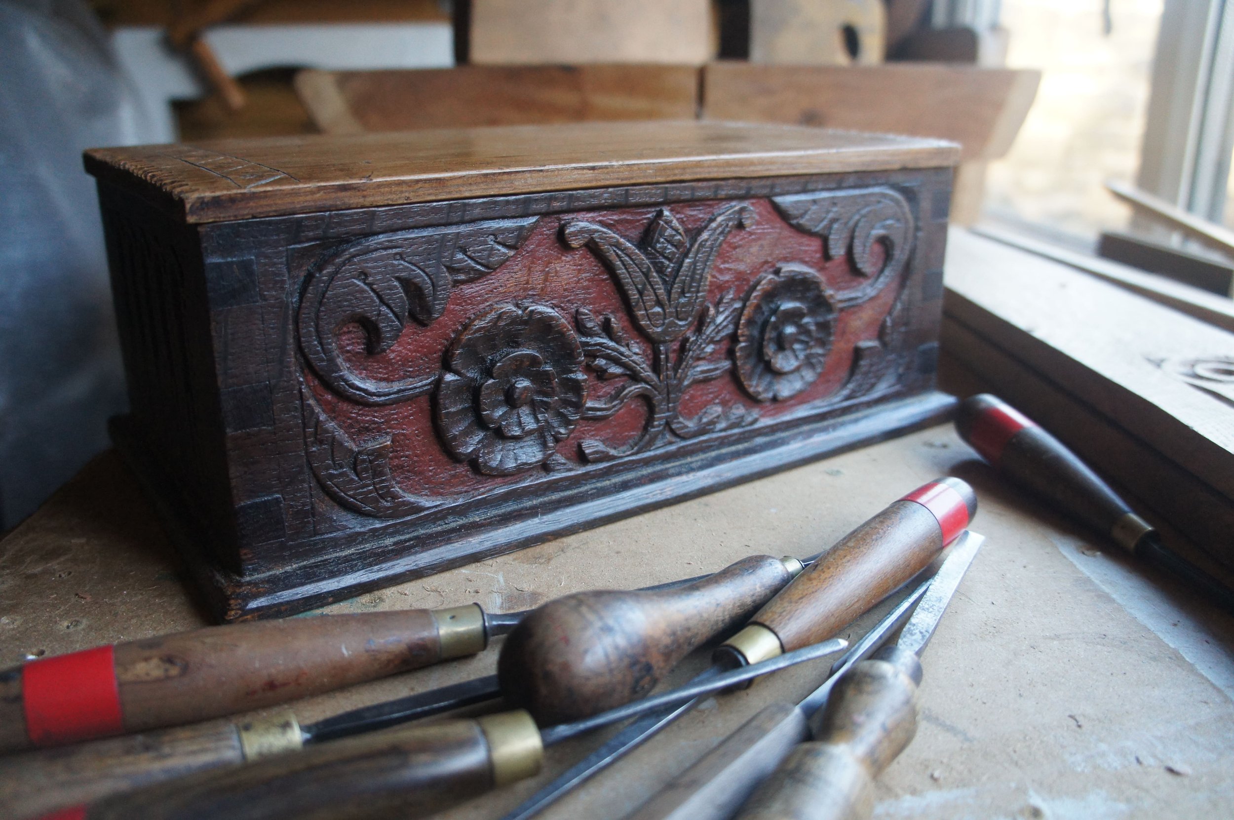 Close-up of a vintage wooden box with ornate carvings, sitting on a workbench with various chisels around it, near a window.