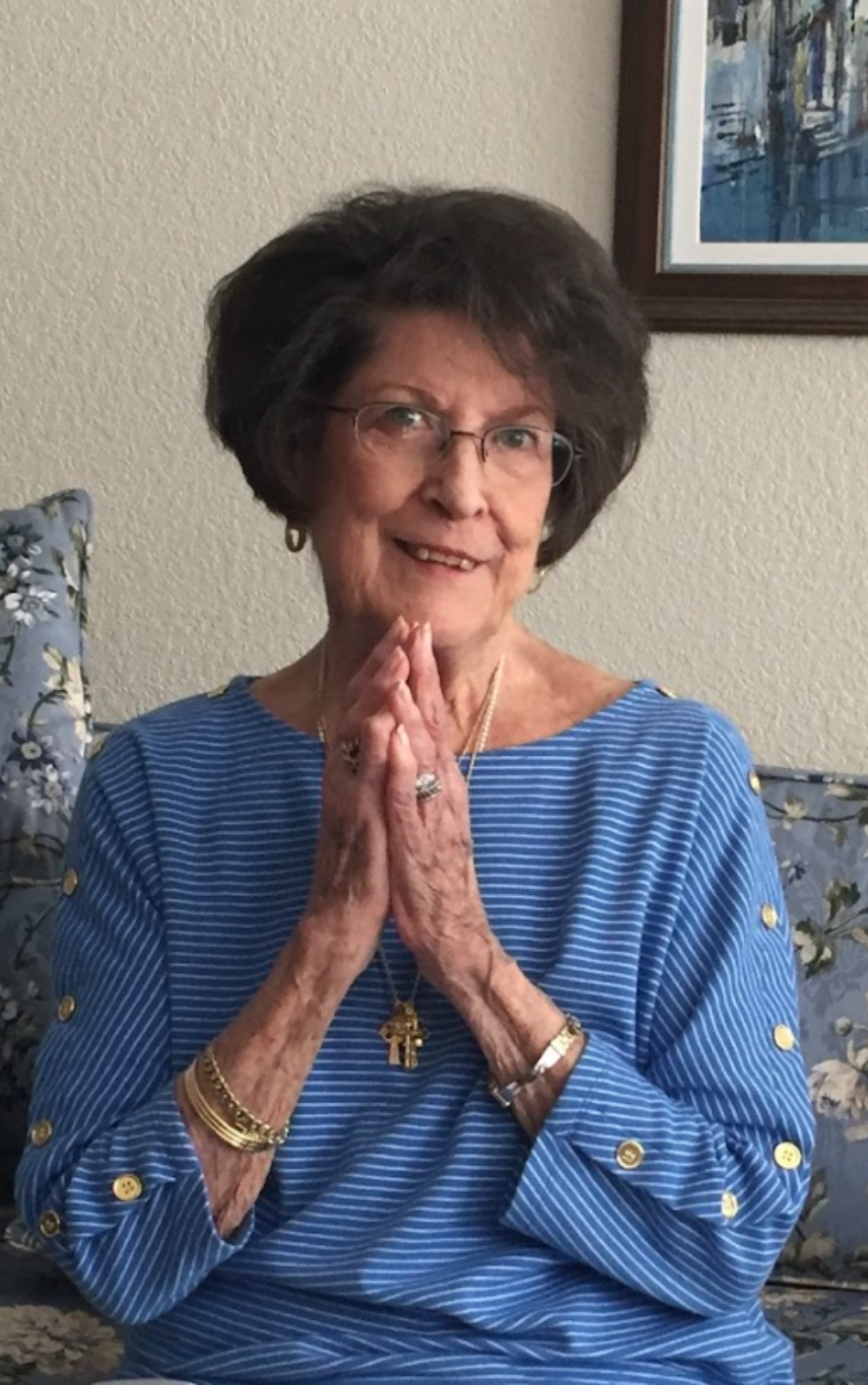 An elderly woman with short gray hair, glasses, and a cheerful expression, sitting on a floral patterned couch with a blue and white striped shirt, clasping her hands in prayer. She is wearing jewelry, including rings, bracelets, and a necklace. A framed painting hangs on the wall behind her.