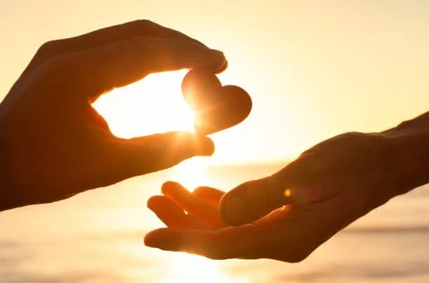 Two hands are shown, one holding the sun between the thumb and index finger, and the other hand underneath, capturing the sunlight during sunset or sunrise over the water.