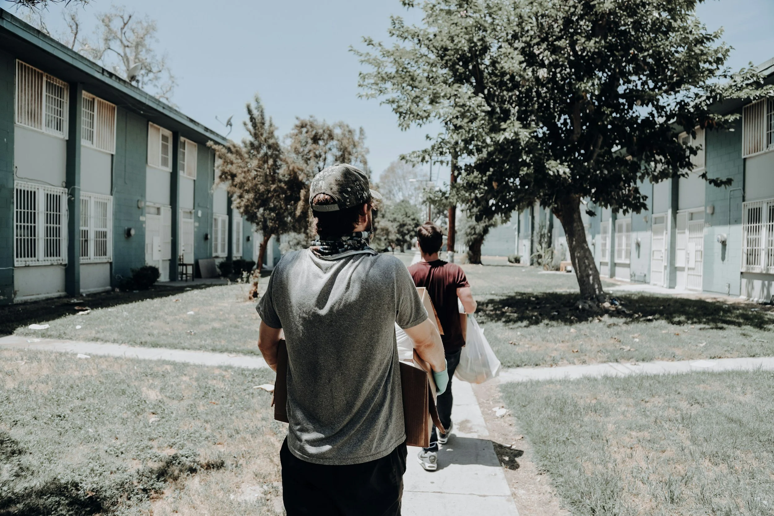 Two young men walking on a sidewalk in a residential neighborhood with apartment buildings on either side and trees overhead.