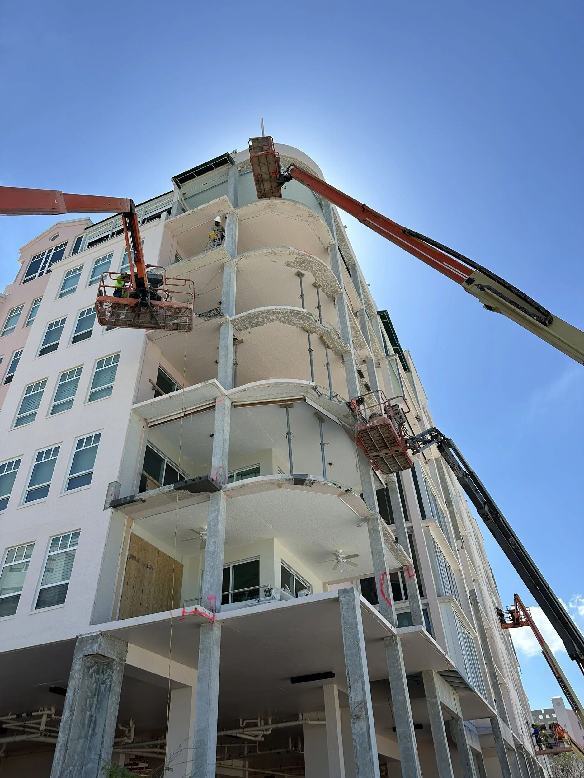 Construction workers on scaffolding and lifts working on the exterior of a multi-story building under construction, with a clear blue sky in the background.