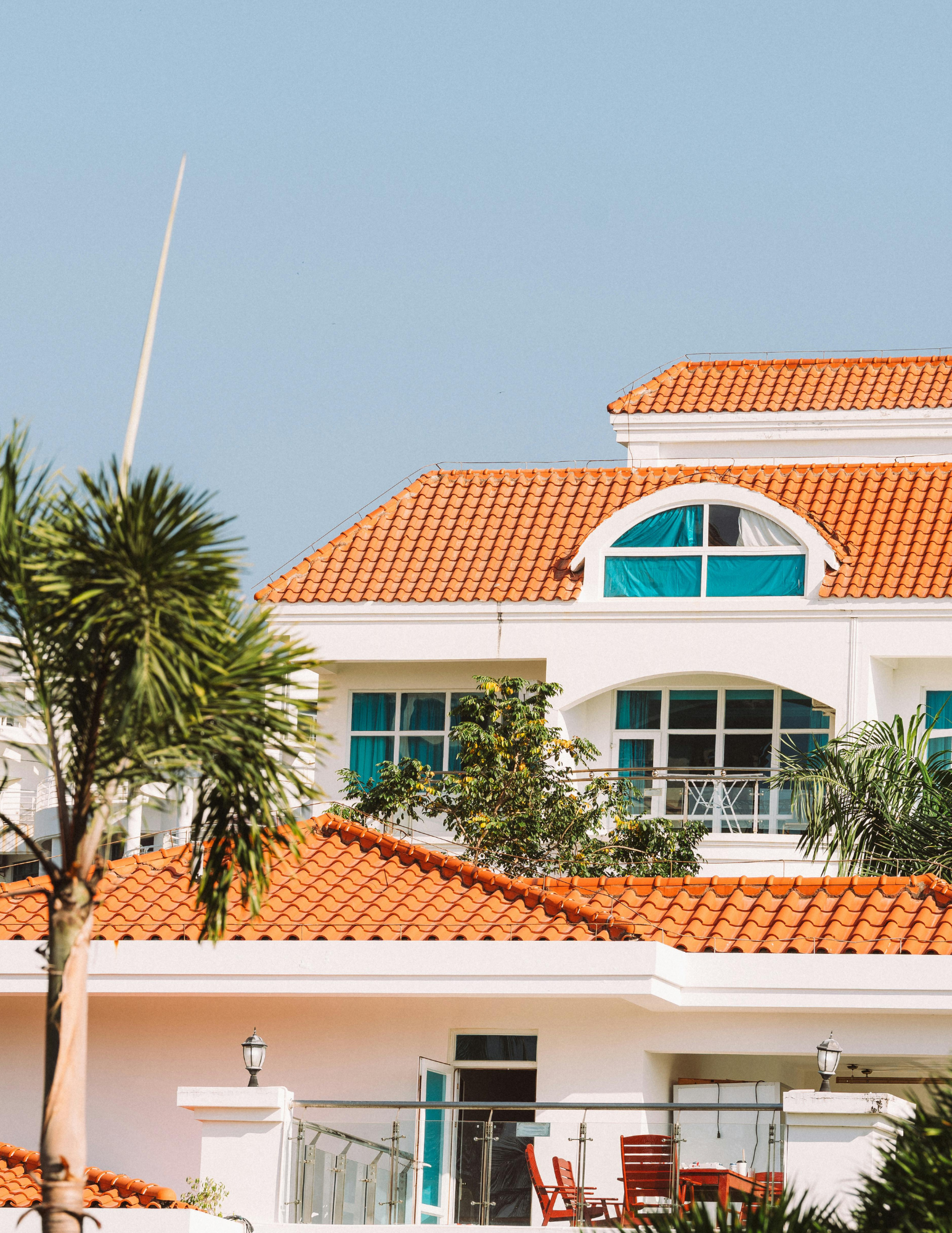 A white multi-story house with red tile roof and large arched windows, outdoor balcony with red chairs, plants, and a palm tree in front.