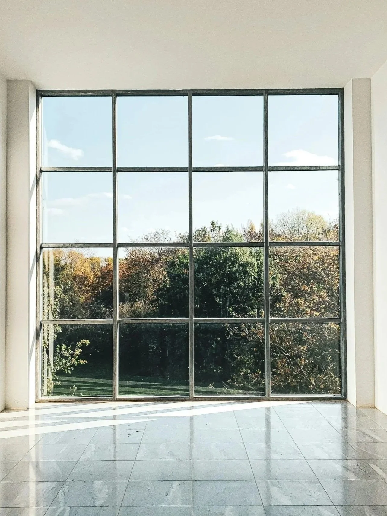 Interior of a building with a large window and tiled floor, showing a view of trees and a blue sky outside.