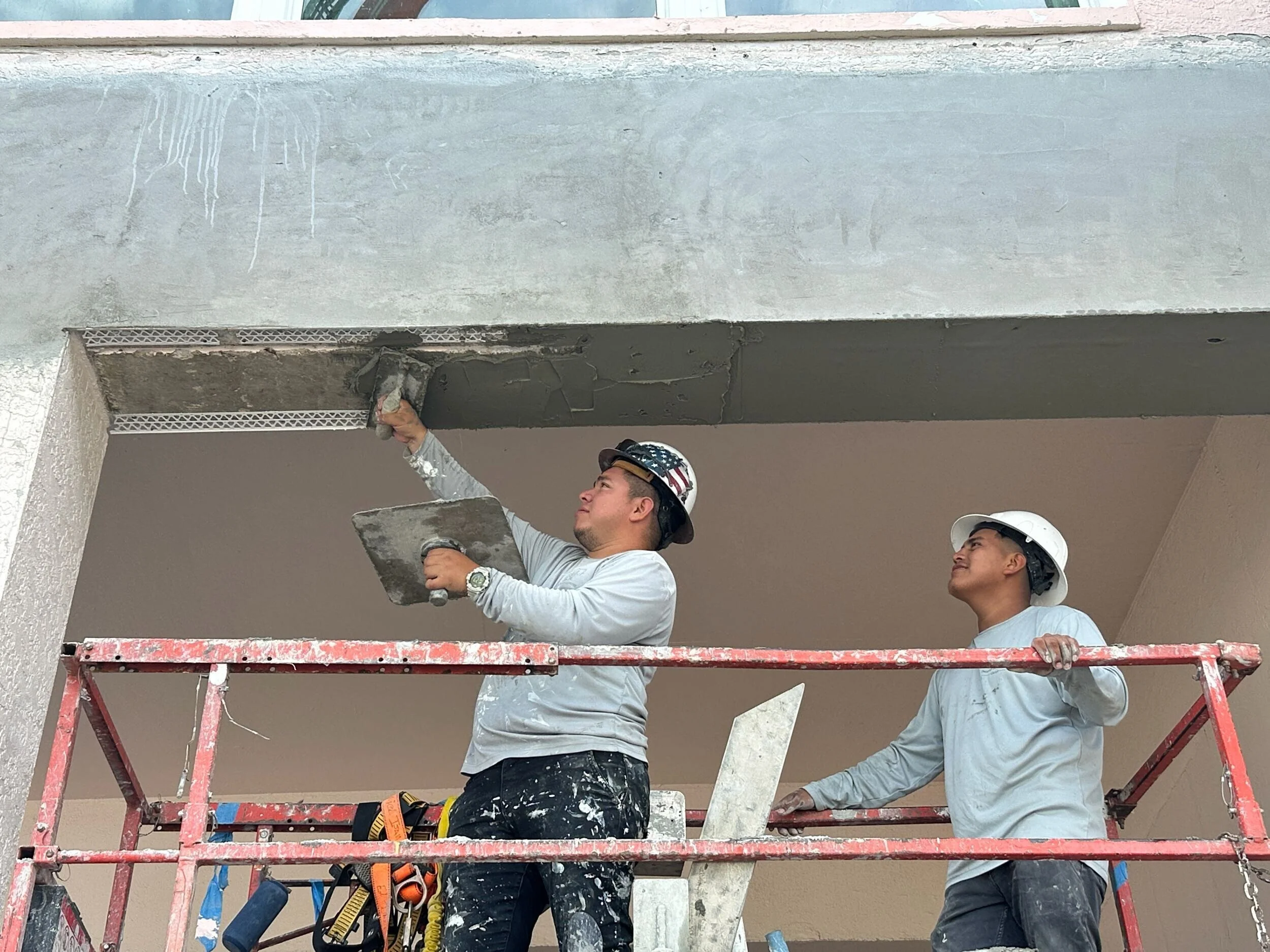 Two construction workers wearing PDG shirts, tool belts, and hard hats, standing on scaffolding and working on installing a concrete ceiling or wall.