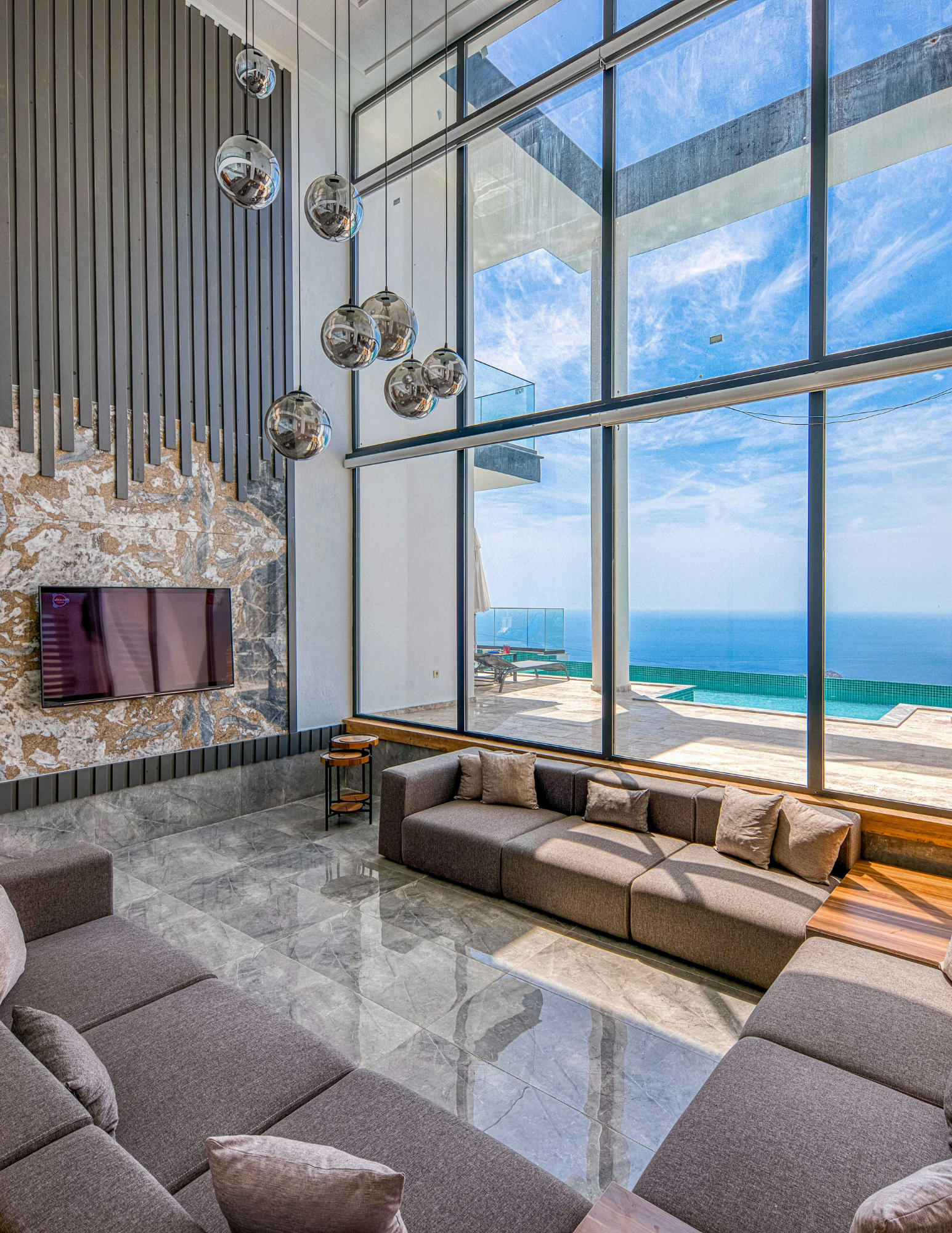Modern living room with large floor-to-ceiling windows overlooking a pool and ocean view, featuring gray sectional sofas, beige pillows, marble flooring, a wall-mounted flat-screen TV, and unique hanging light fixtures with spherical reflective shades.