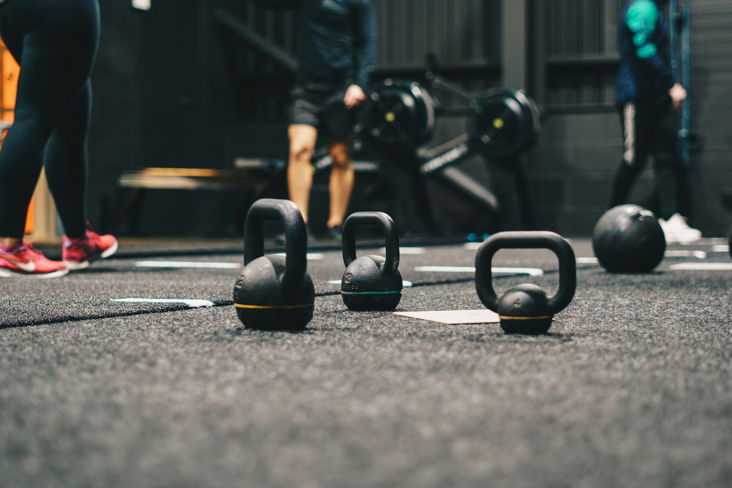 Kettlebells and a medicine ball on the gym floor with people working out in the background.
