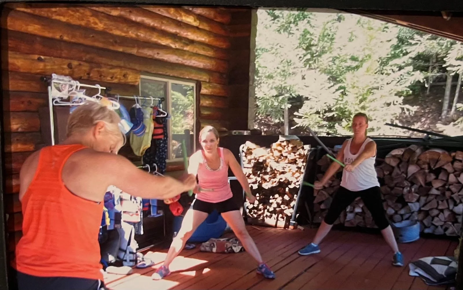 Three women exercising with resistance bands on a wooden porch surrounded by wood stacked for a firewood pile, with trees visible in the background.