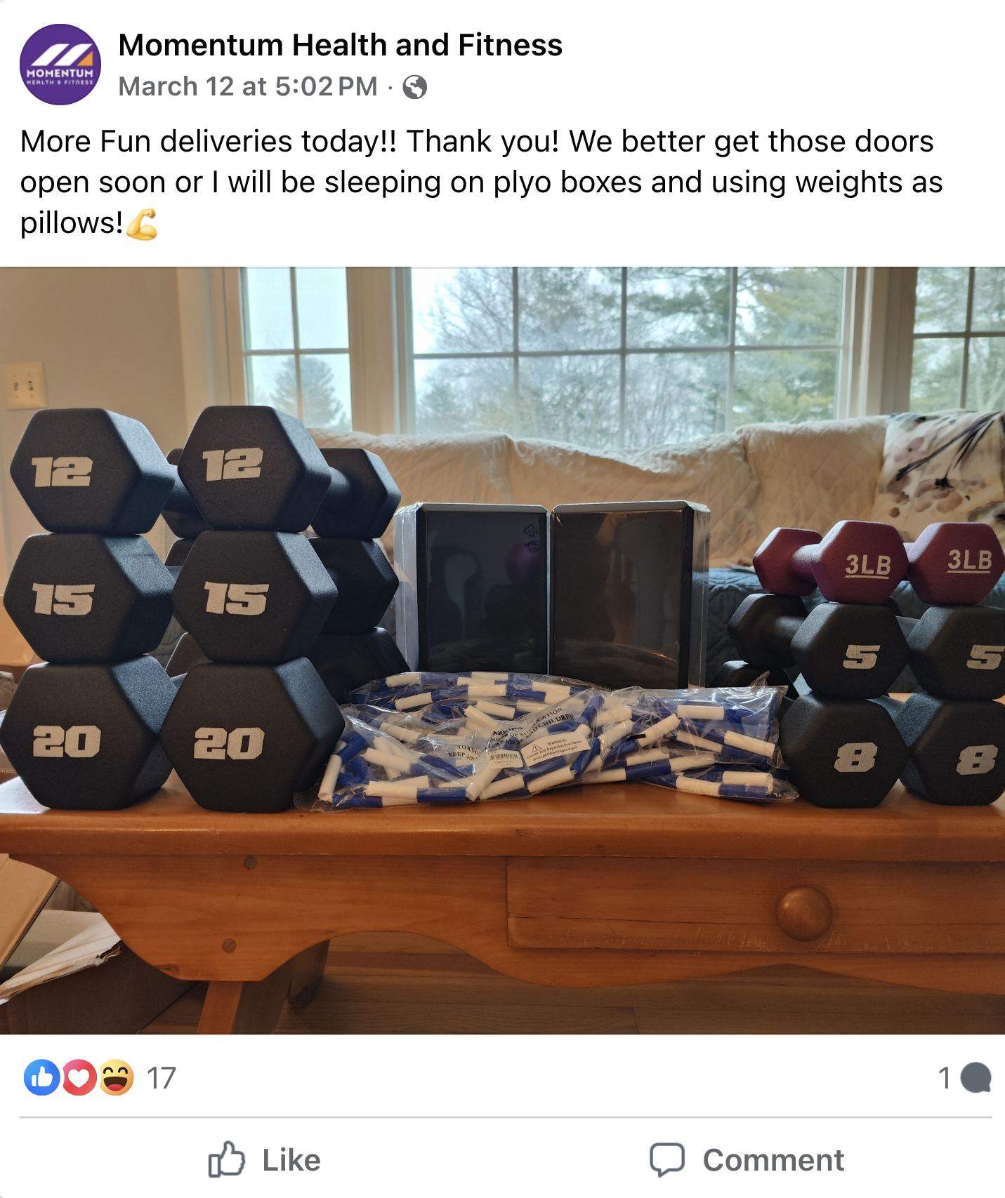Set of black and red dumbbells of various weights, stacked on a wooden table along with packages of chalk and two black boxes, with a background of a couch and windows showing trees outside.
