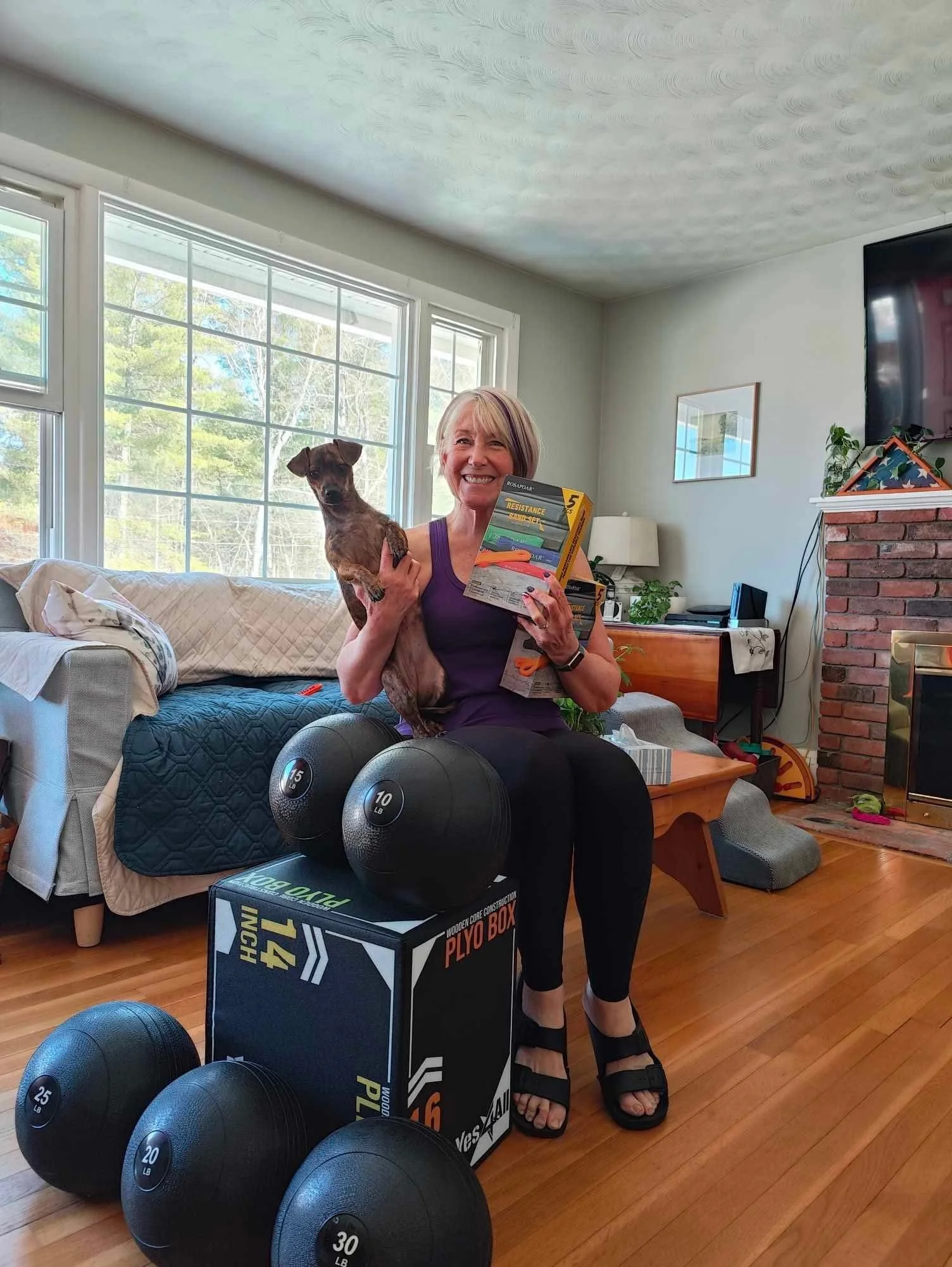 A woman sitting on a chair inside a living room, smiling and holding a small dog in her left arm and a box of resistance bands in her right hand. In front of her on the floor are three black exercise balls labeled 15 lb, 20 lb, and 25 lb. The room has large windows letting in natural light, a couch with pillows and a quilt, a fireplace, and a television.