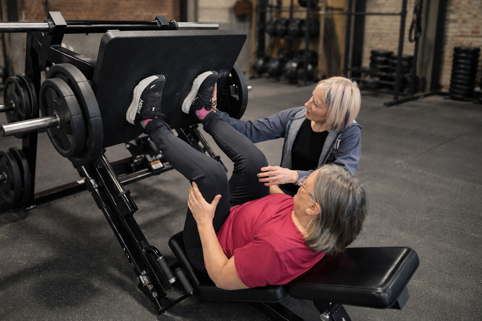 An elderly woman lying on a leg press machine with her legs extended, guided by a trainer helping her perform a workout at the gym.
