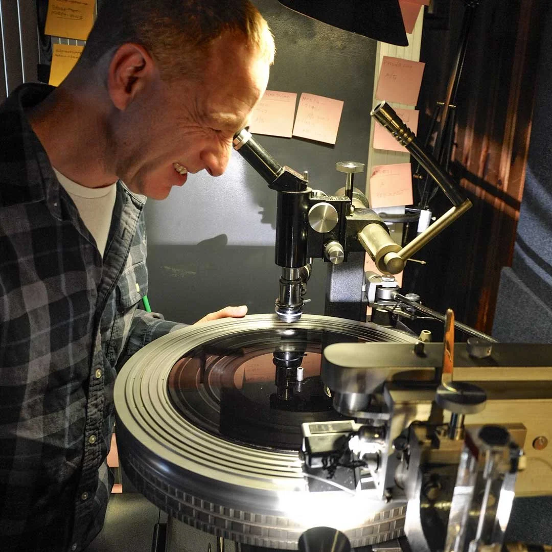 A man with a short haircut wearing a plaid shirt looking through a microscope at a spinning record on a turntable.