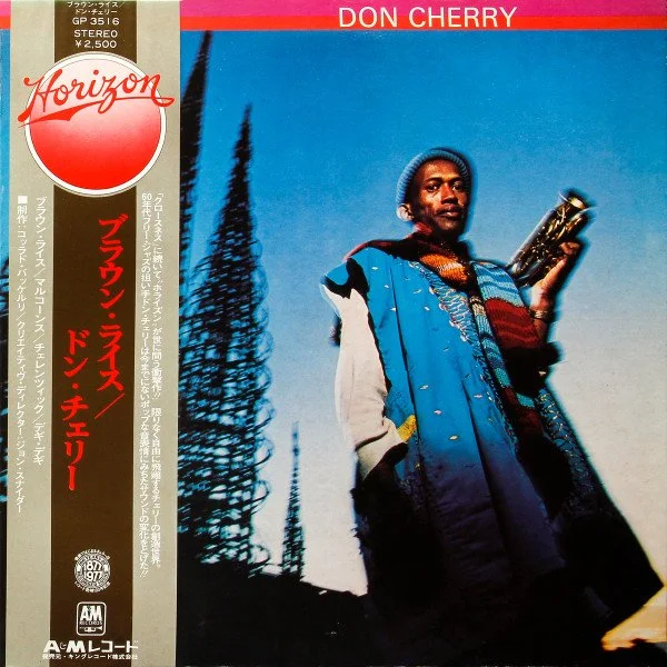 Album cover for Don Cherry titled "Brown Rice" featuring a man outdoors holding a saxophone, wearing layered clothing and a hat, with tall trees and a clear blue sky in the background.