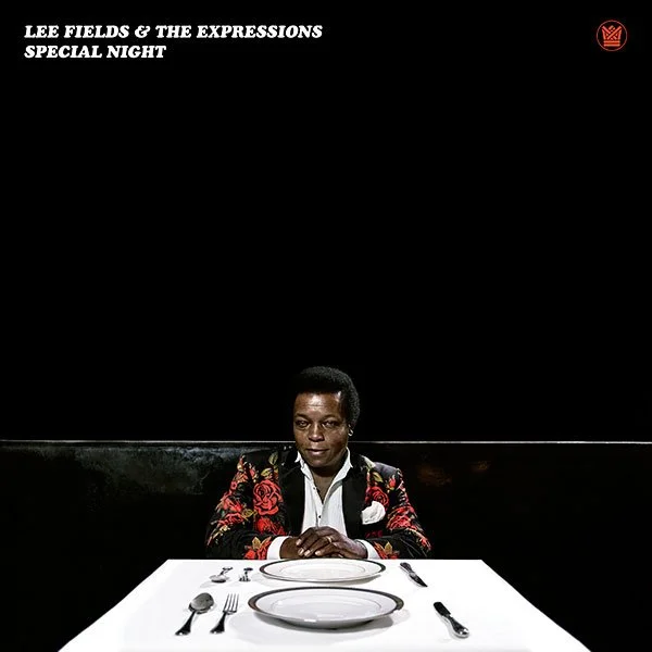 Lee Fields sitting alone at a restaurant table with a black background, empty plates, and silverware, in front of a black wall with white text and a red logo.