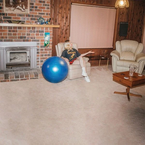 A young girl sitting on a beige sofa in a living room, with a blue exercise ball beside her, brick fireplace, wooden paneling, and a window with a white shade in the background.