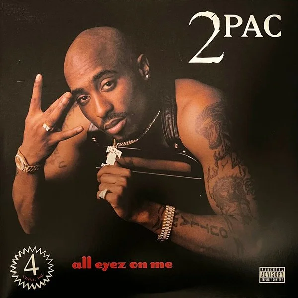 Close-up image of a man with a shaved head, tattoos on his arms, wearing jewelry, making a hand gesture, and looking at the camera. Text includes "2 PAC," "all eyez on me," and a parental advisory label.