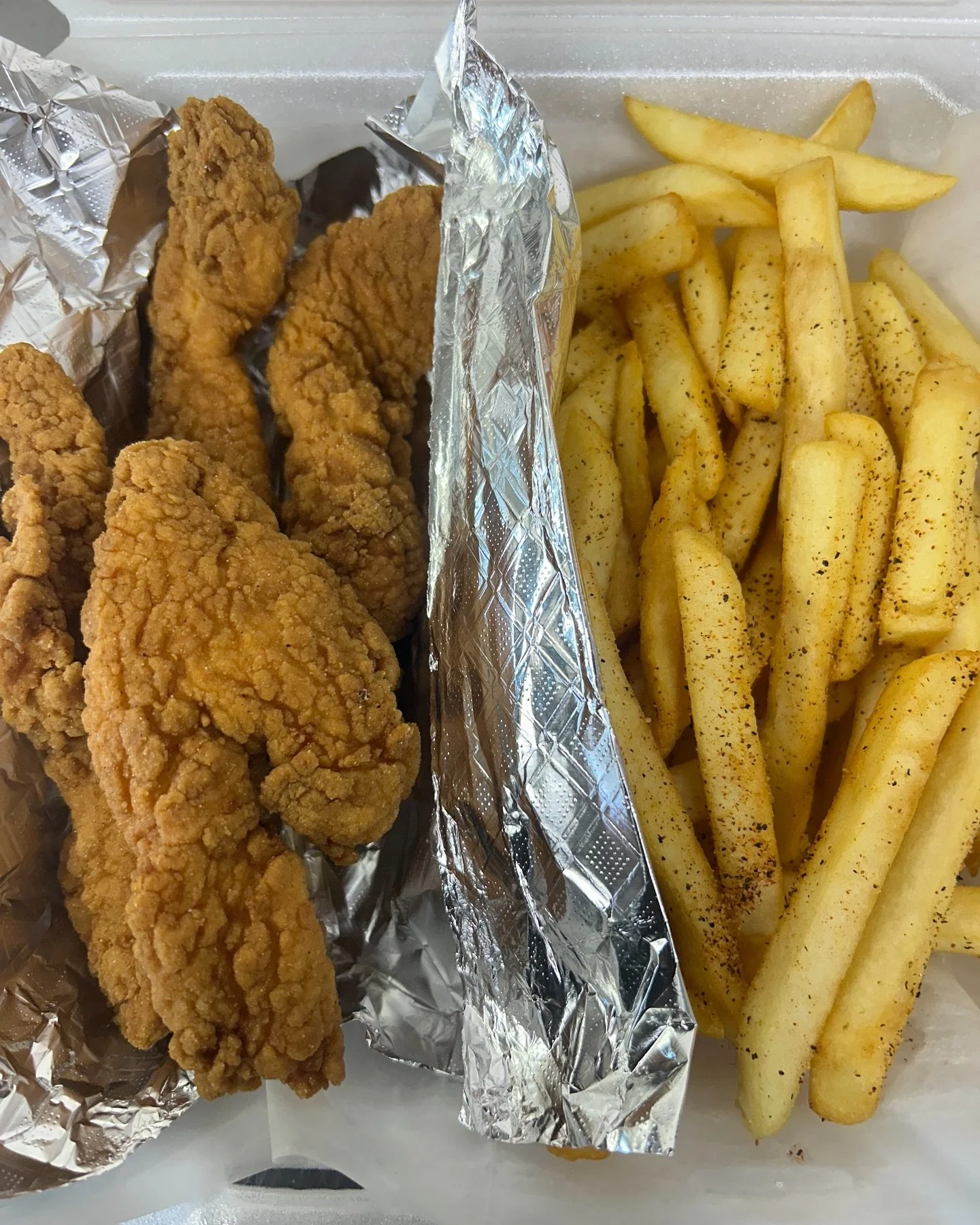 Fried chicken tenders with French fries seasoned with black pepper, served in a foil container.