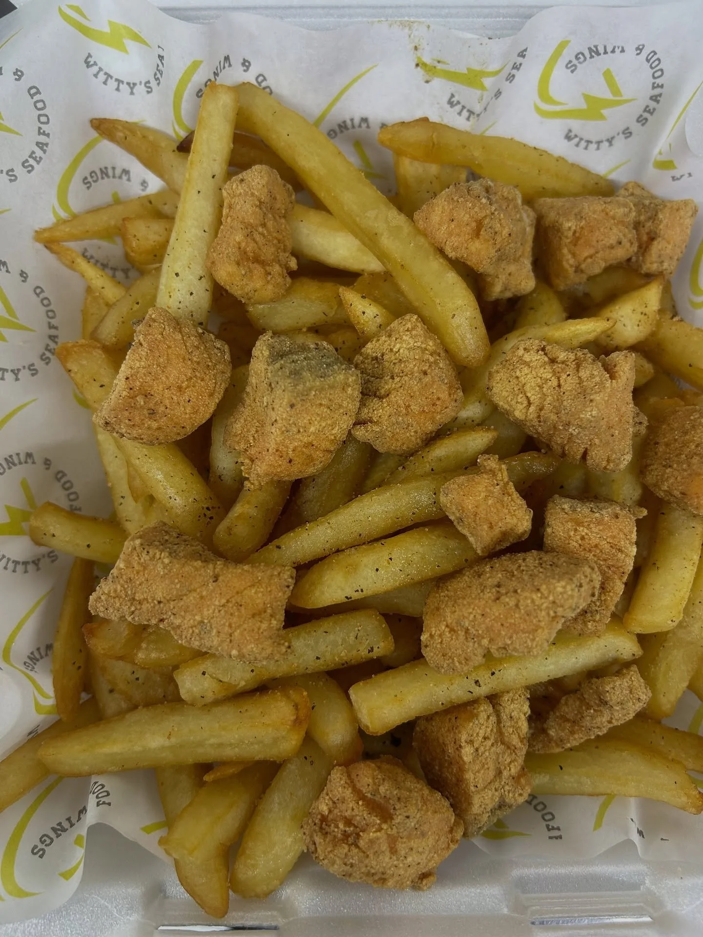 French fries with pieces of fried chicken on top, served on branded paper.