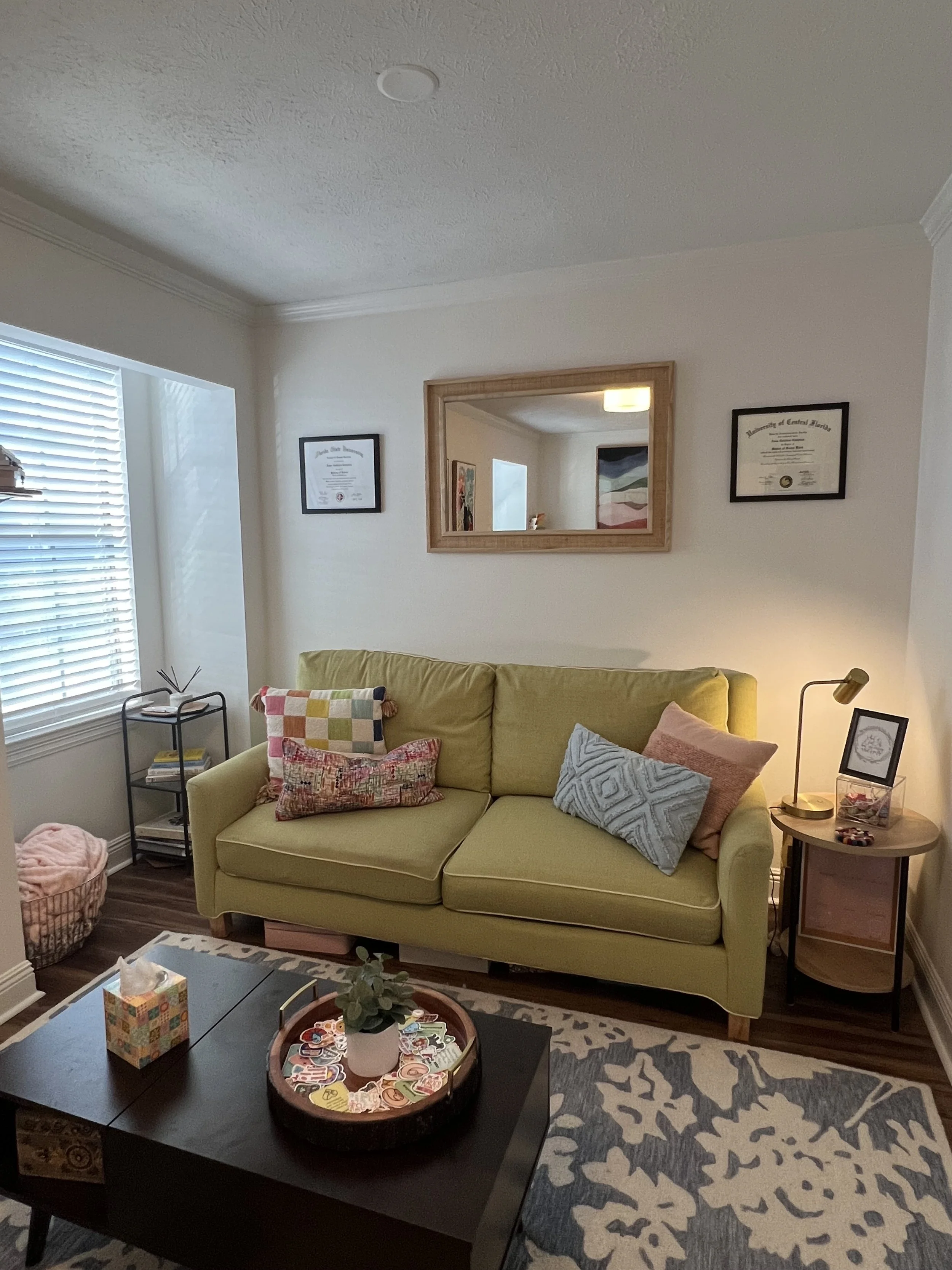 Living room with green sofa, pillows, coffee table with plant and decorative items, window with blinds, wall mirror, framed diploma, and art on the walls.