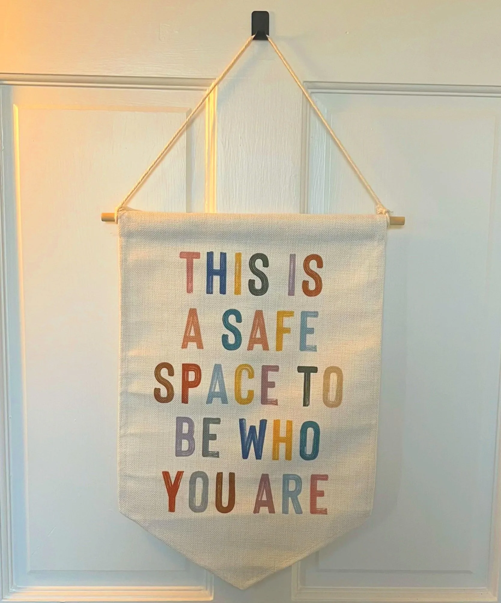 A fabric wall hanging with the message 'This is a safe space to be who you are' in colorful letters hanging on a door.
