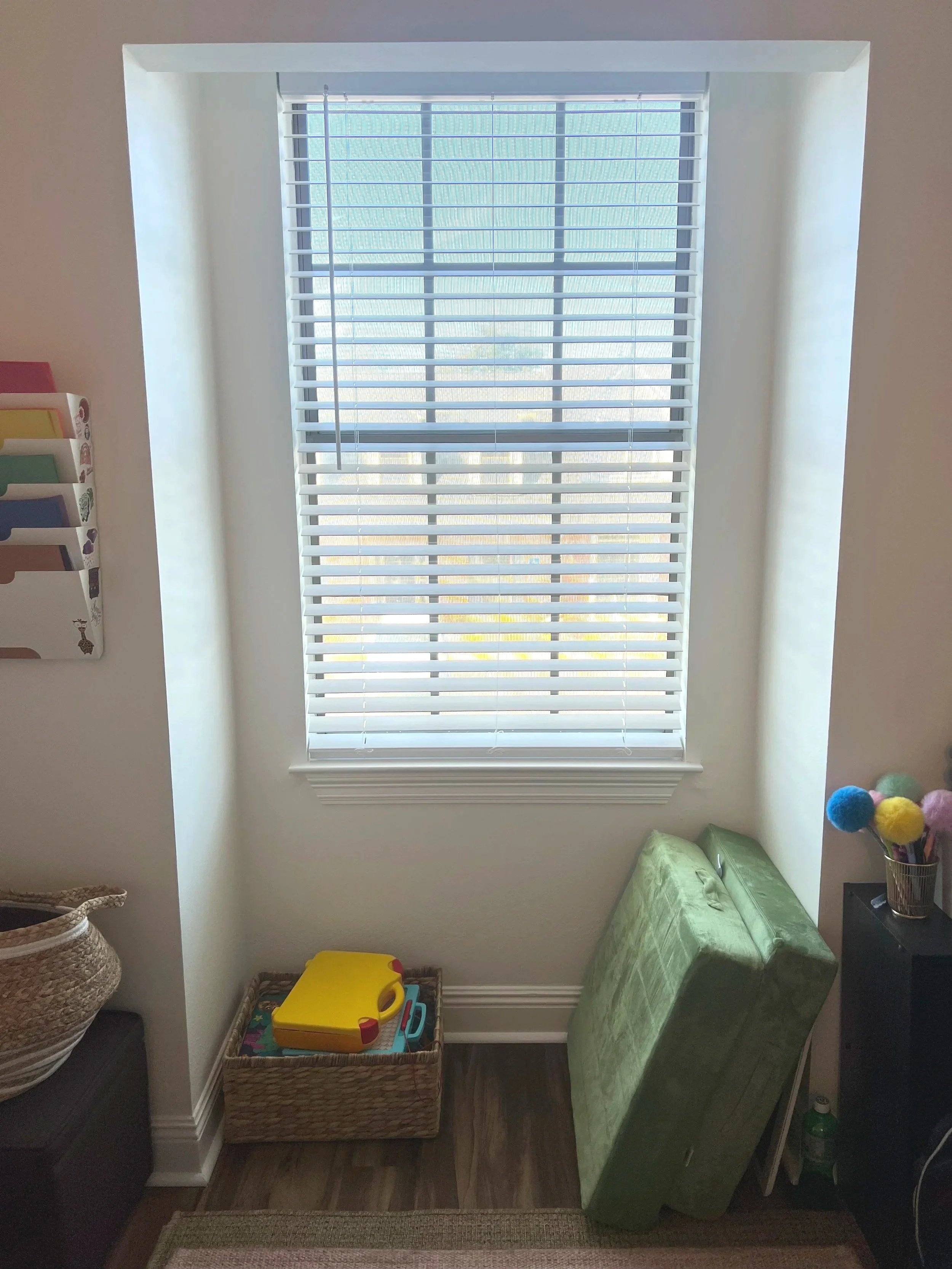 A small, sunlit nook with a large window with white horizontal blinds, a green folding mattress leaning against the wall, a black side table with a jar of colorful pom-pom pens, and a woven basket with a yellow toy on top.