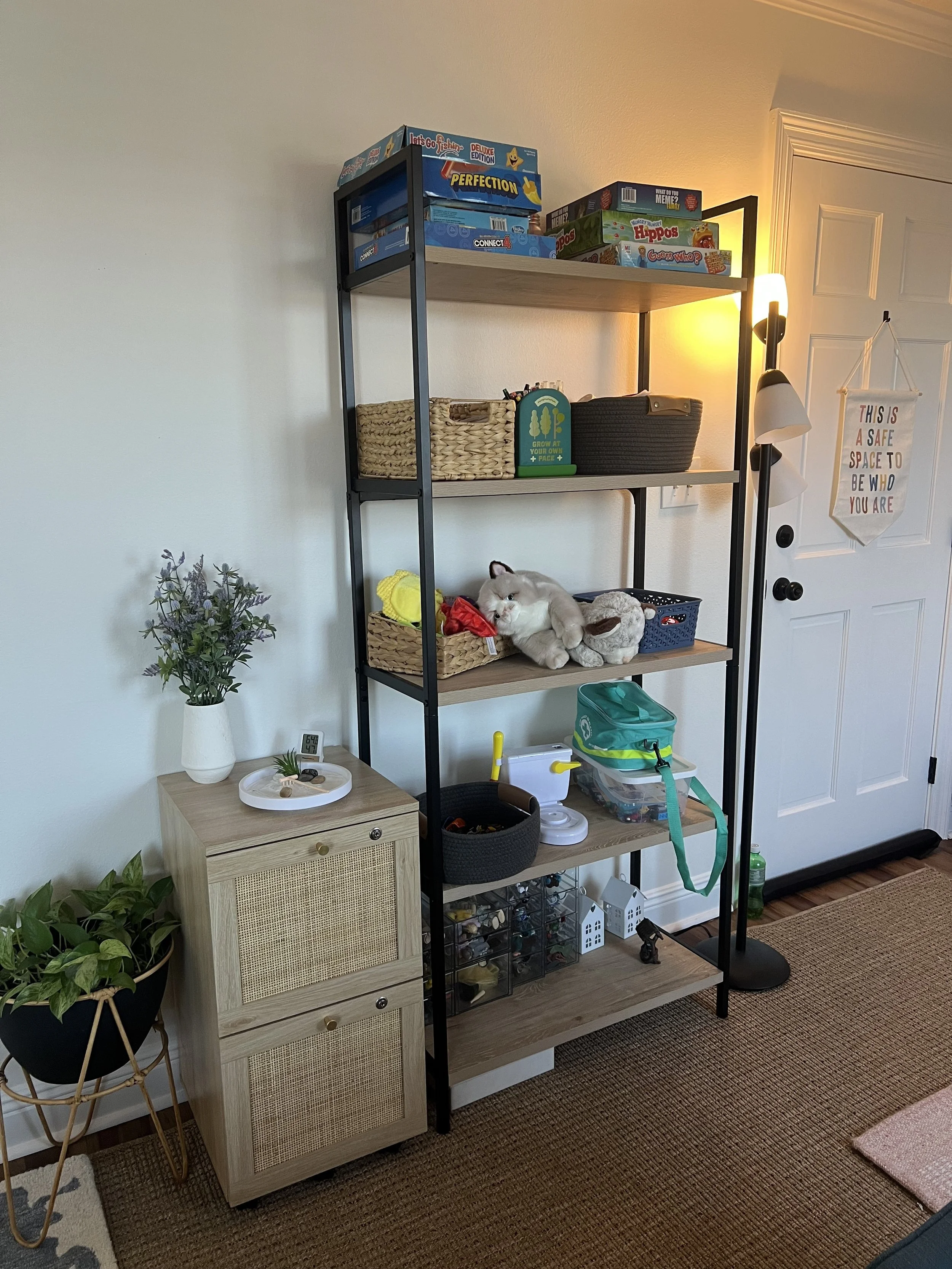 A black and wooden shelving unit with board games on top and stuffed animals on middle shelves, next to a small wooden cabinet with a plant and clock on top, in a living room corner.
