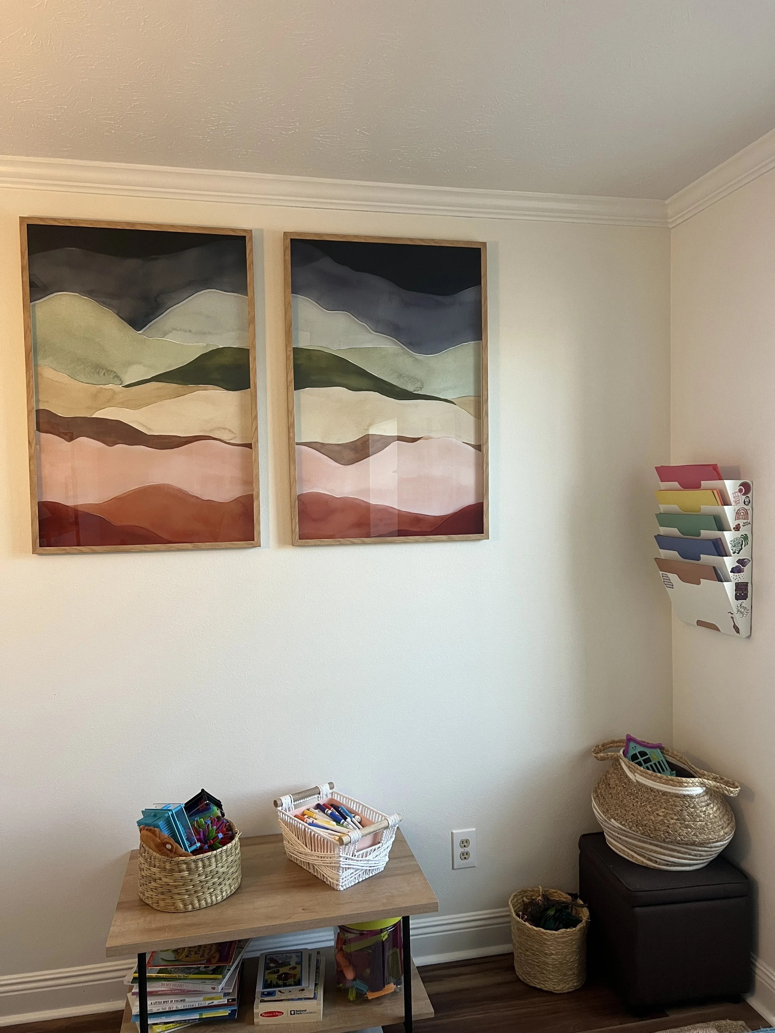 A children's play area with two framed landscape paintings on the wall, a small wooden table with baskets of coloring supplies, and a wall-mounted organizer with color-coded folders.