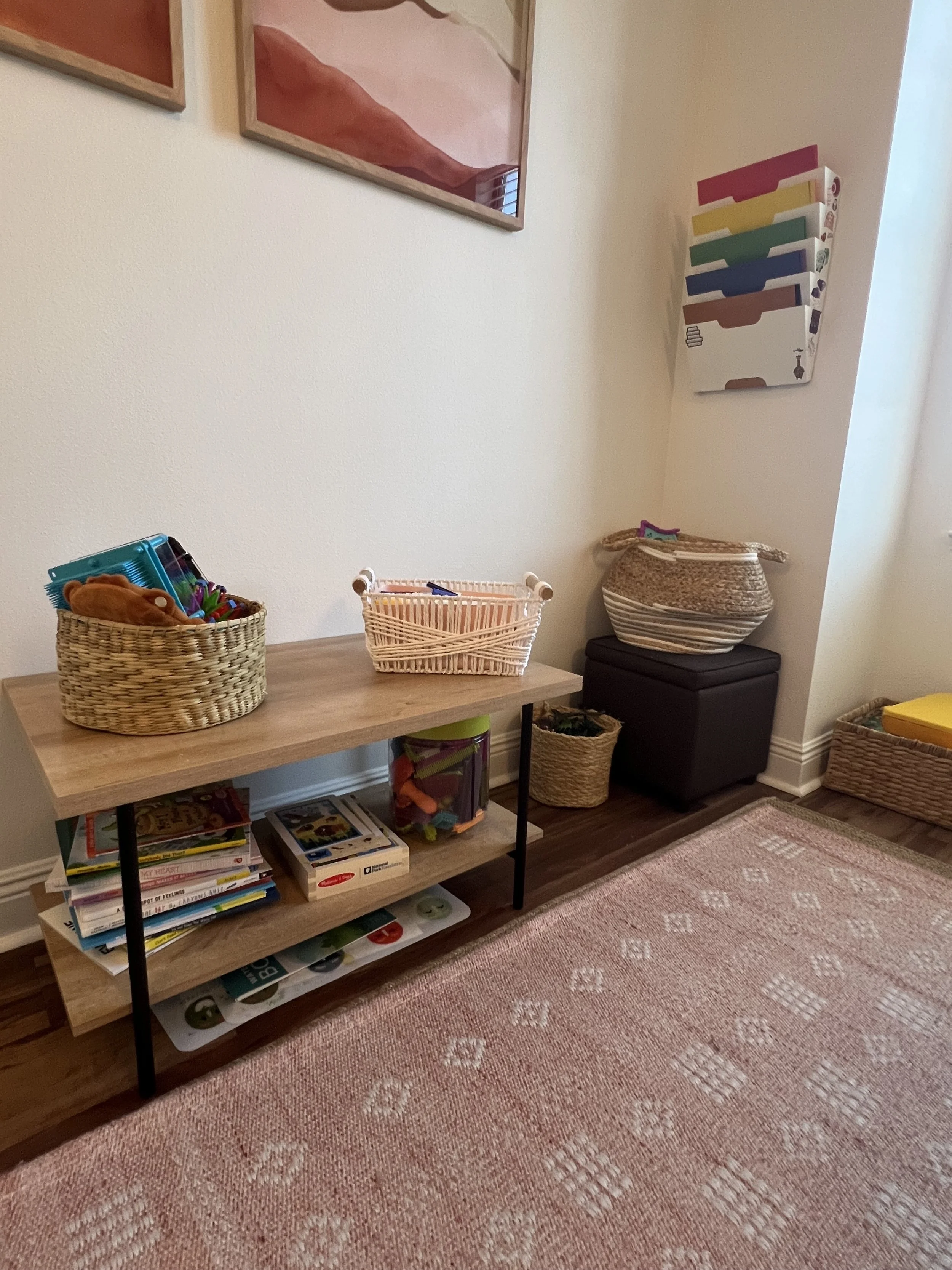 A small wooden table with black metal legs holding baskets of toys, books, and games, positioned in a children's playroom corner with wall art and colorful storage bins.