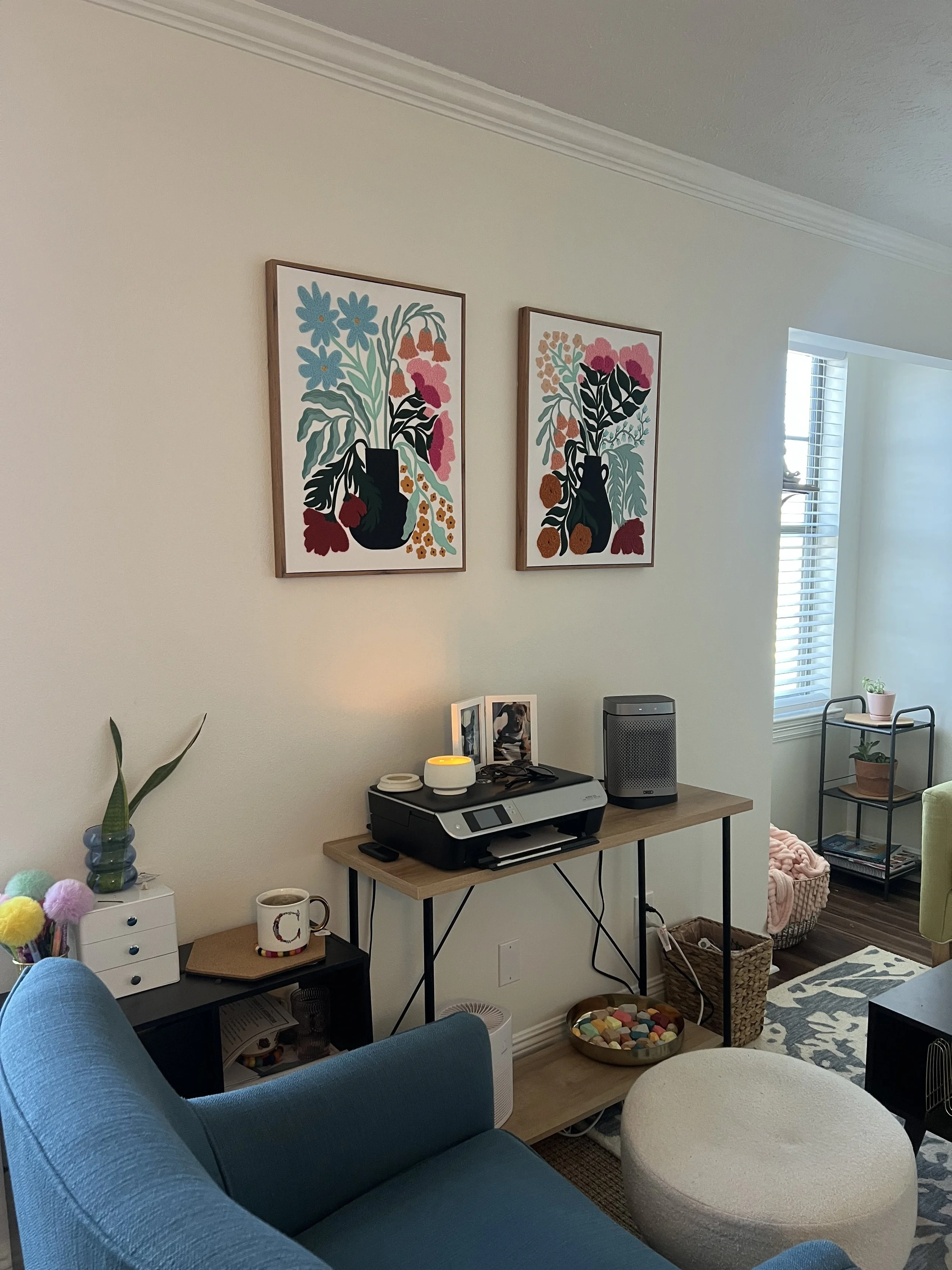 Living room with two colorful floral paintings on the wall above a wooden table holding a printer, framed photographs, an essential oil diffuser, and a speaker. There are various furnishings, including a blue armchair, a small black side table with a