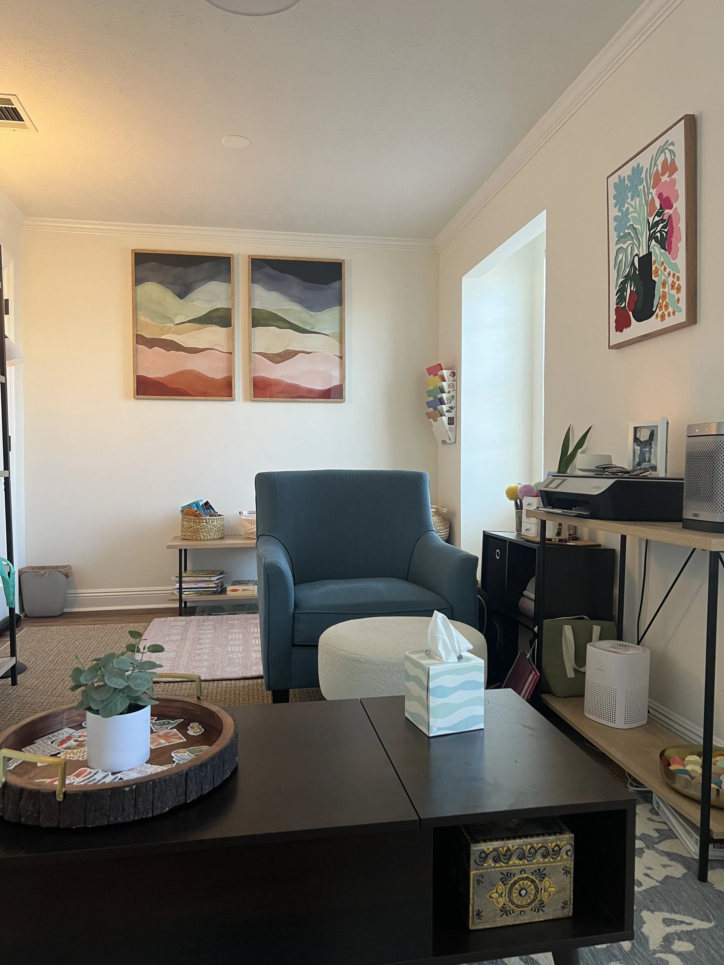 Living room with a blue armchair, paintings on the wall, and a table with a tissue box and a plant.