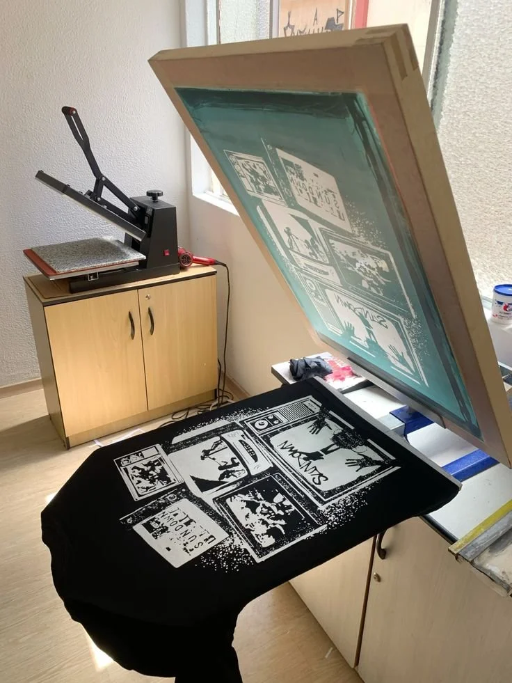 A printing press screen printing a black and white design onto a black fabric, with the printed design visible on the fabric, and the screen positioned above it, ready for printing.