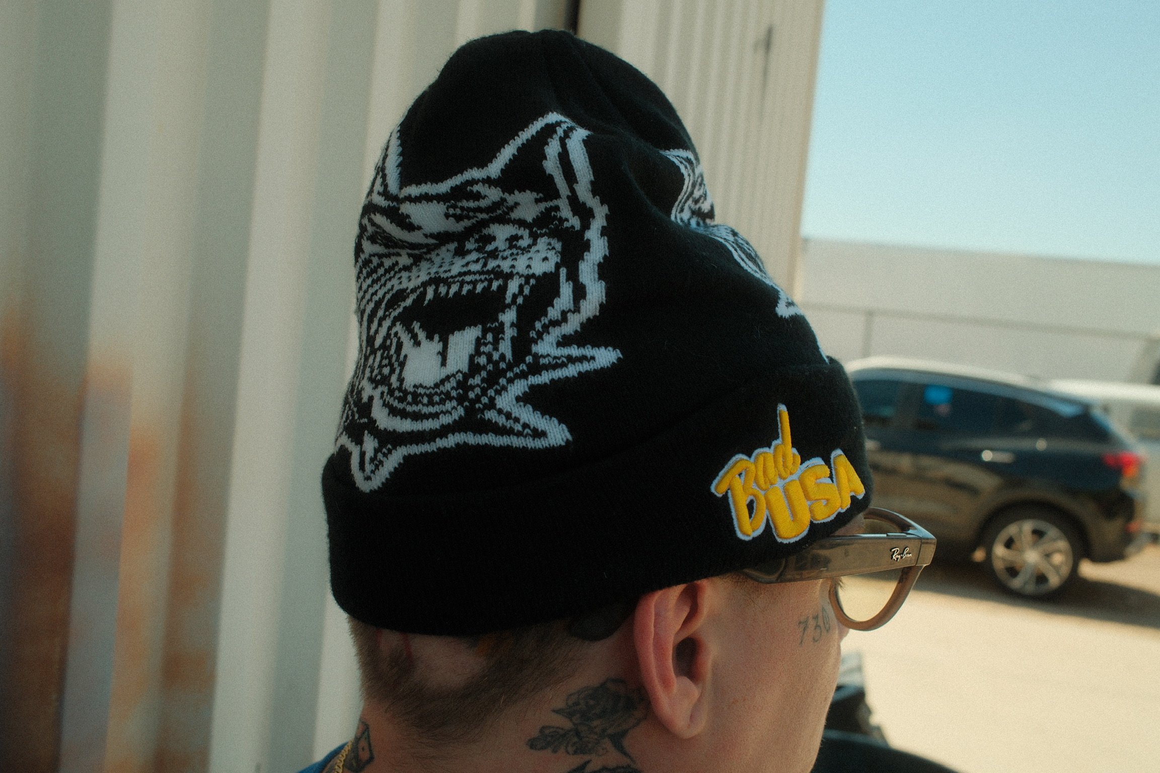 A person wearing a black beanie with a white tiger design and the words 'True BUSA' in yellow and white embroidery, sunglasses, and tattoos on their neck, standing outdoors with cars and a building in the background.